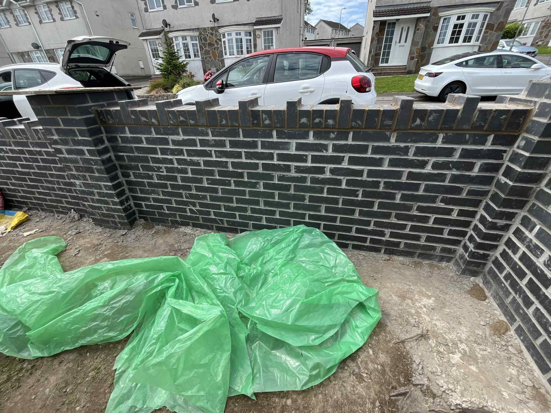 Brick wall Black brick wall under construction with green tarp in front, cars and houses in background.