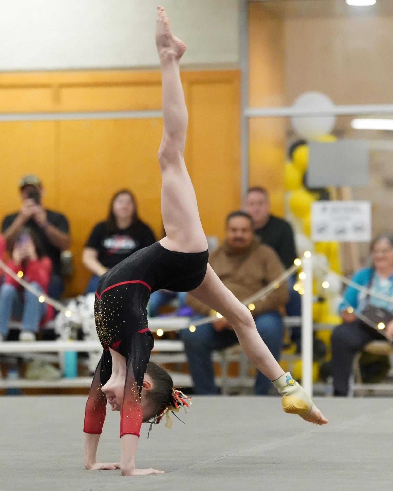Gymnast in black leotard doing a handstand on the floor, with one leg extended up and the other at an angle. Spectators in the background.