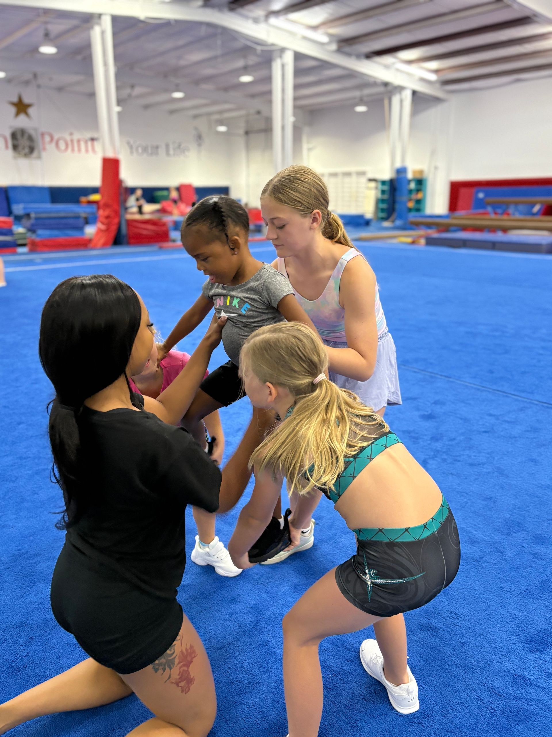 A woman is kneeling down with three young girls on a blue carpet in a gym.