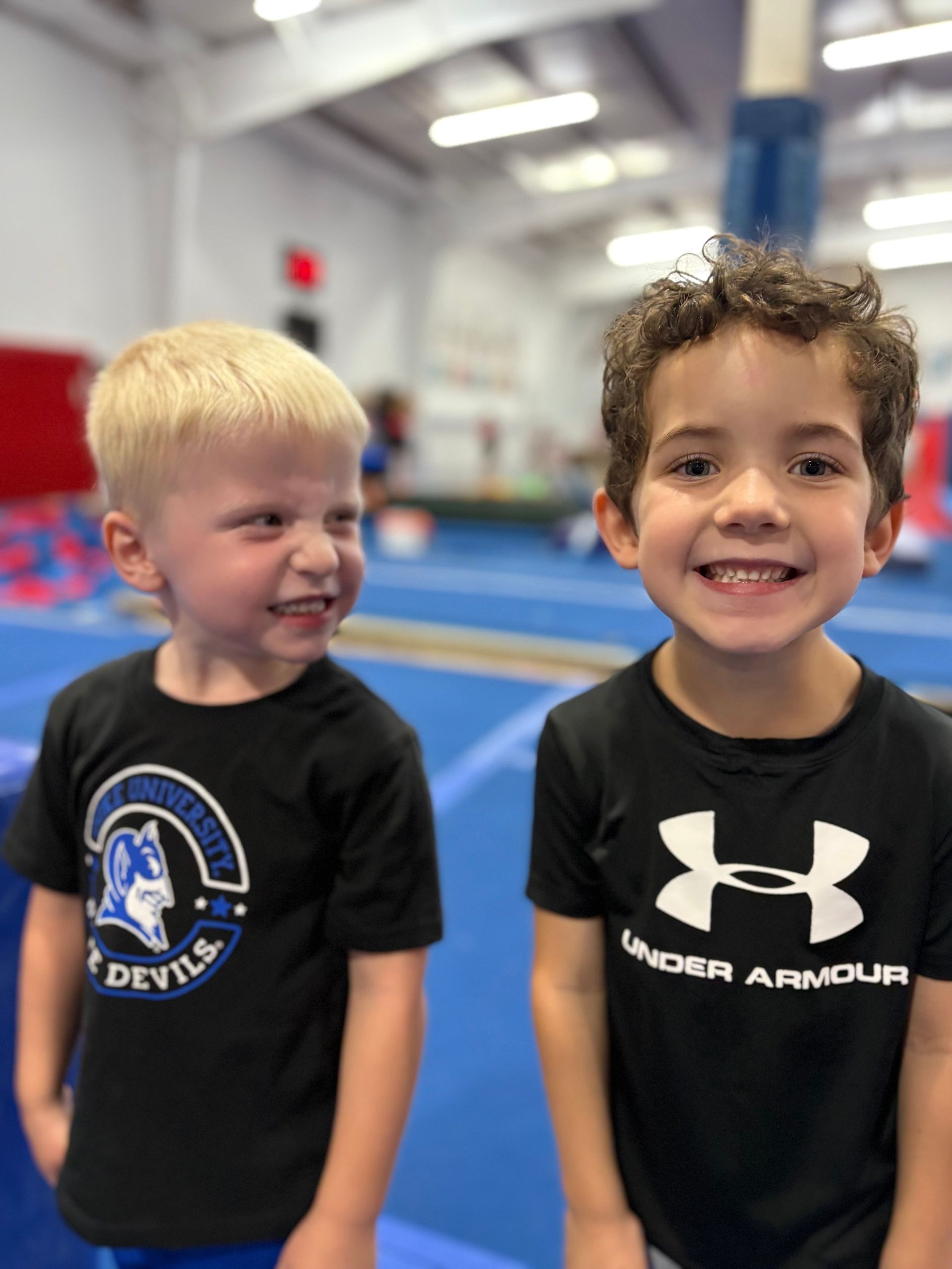 Two young boys smiling at a gymnastics gym; one blond, one with curly hair; wearing black shirts.