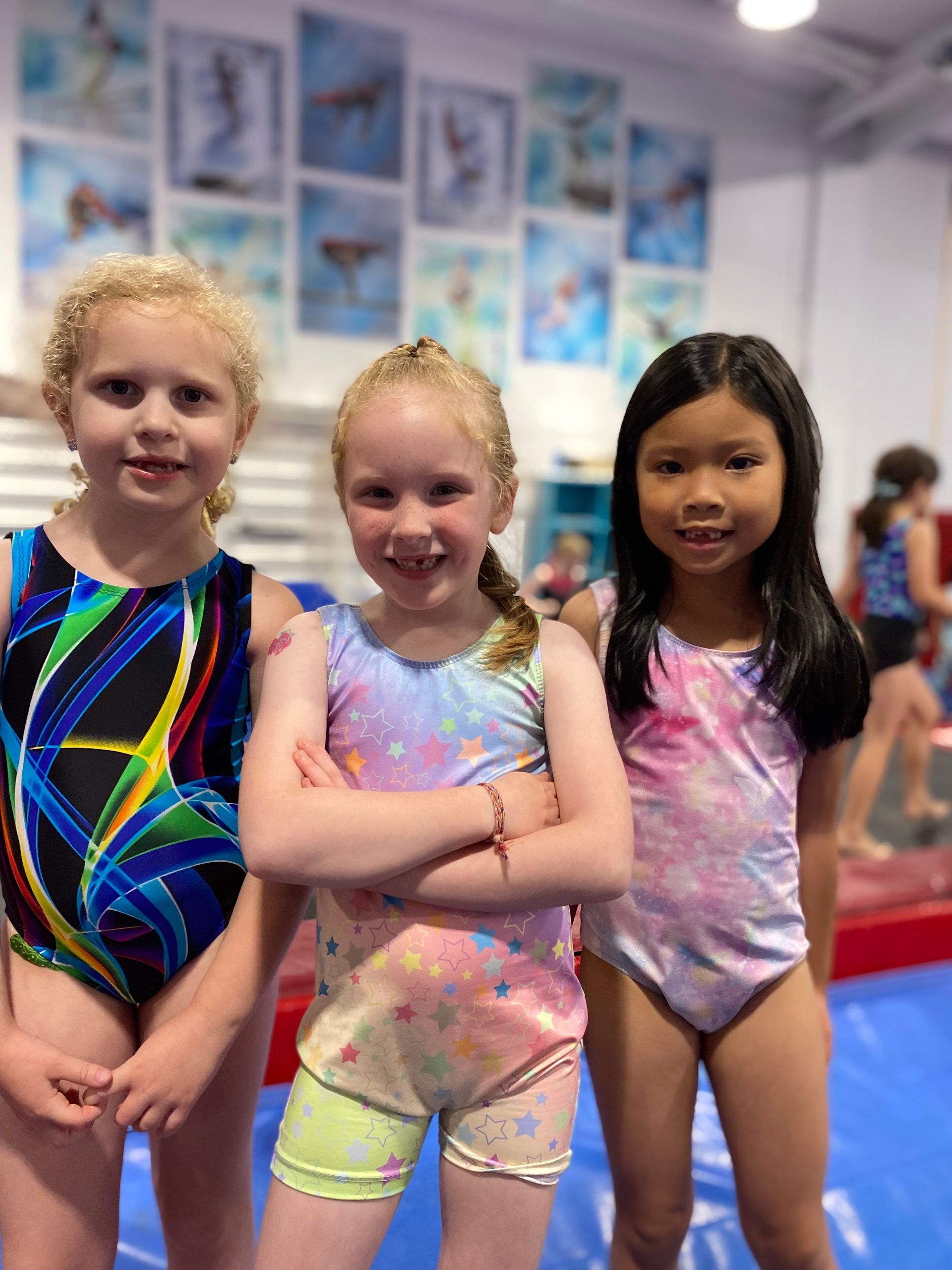 Three young gymnasts in colorful leotards smile at the camera in a gym. The center girl has crossed arms.