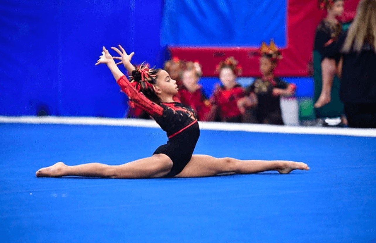 Young gymnast in black and red leotard performs a split on a blue mat, arms raised. Other gymnasts in the background.