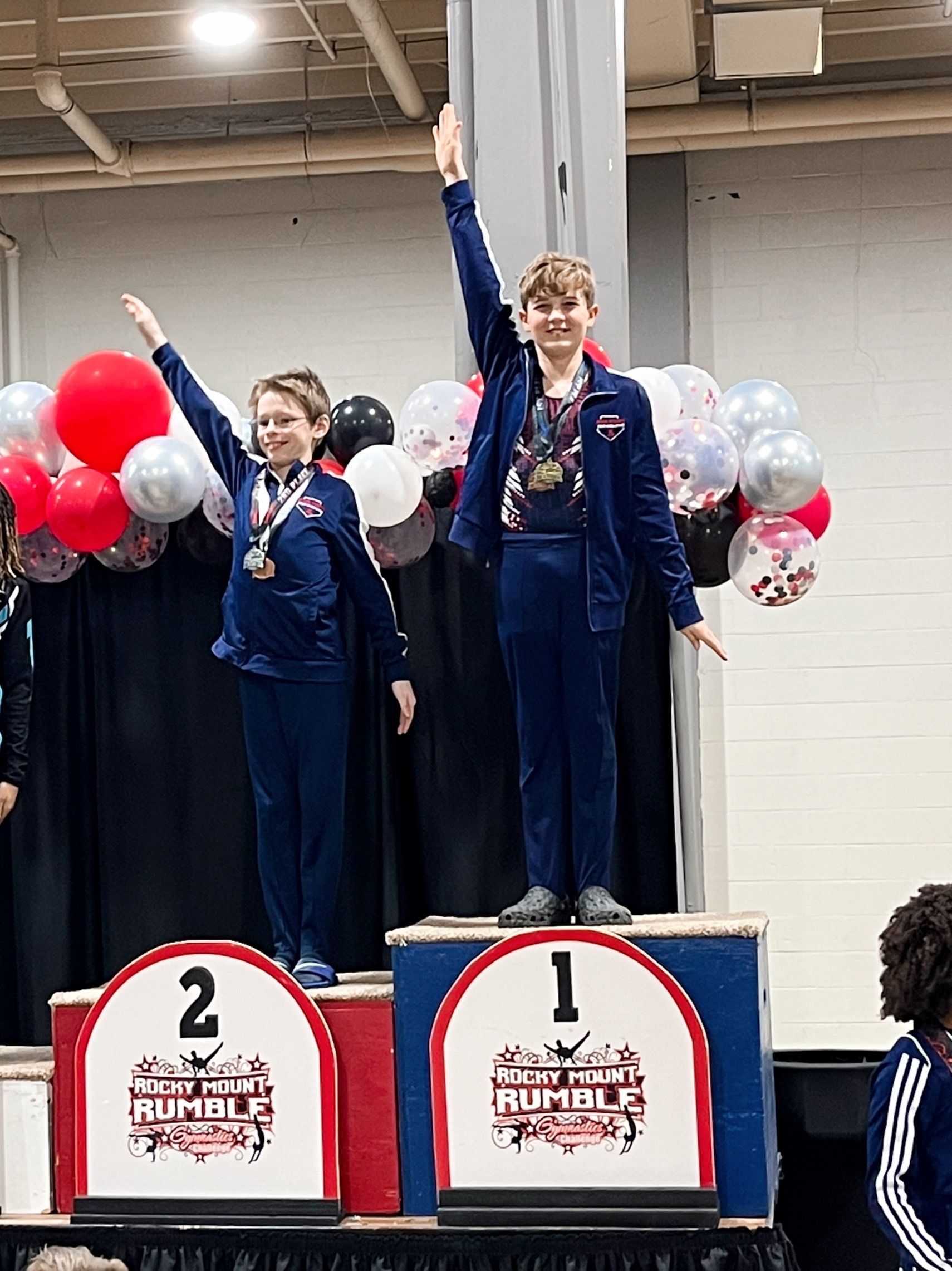 Two young boys on a podium, wearing matching blue suits, raise their arms to celebrate winning medals at a gymnastics competition.