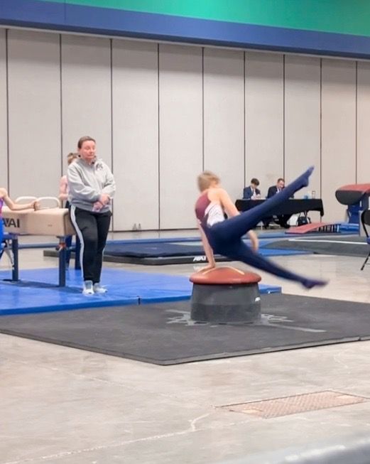 Young gymnast in black and red leotard performs a split on a blue mat, arms raised. Other gymnasts in the background.