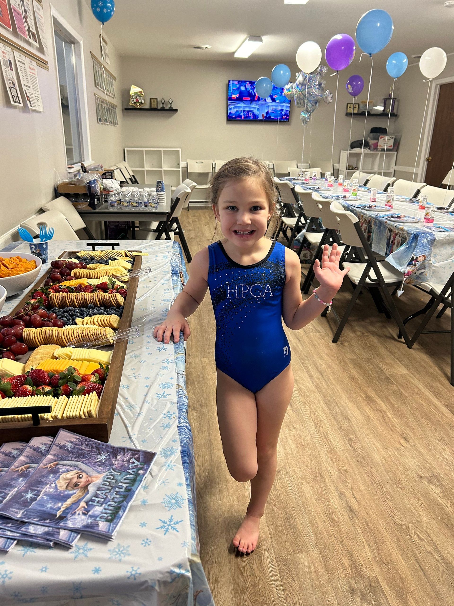 A little girl in a blue leotard is standing in a room with tables and balloons.