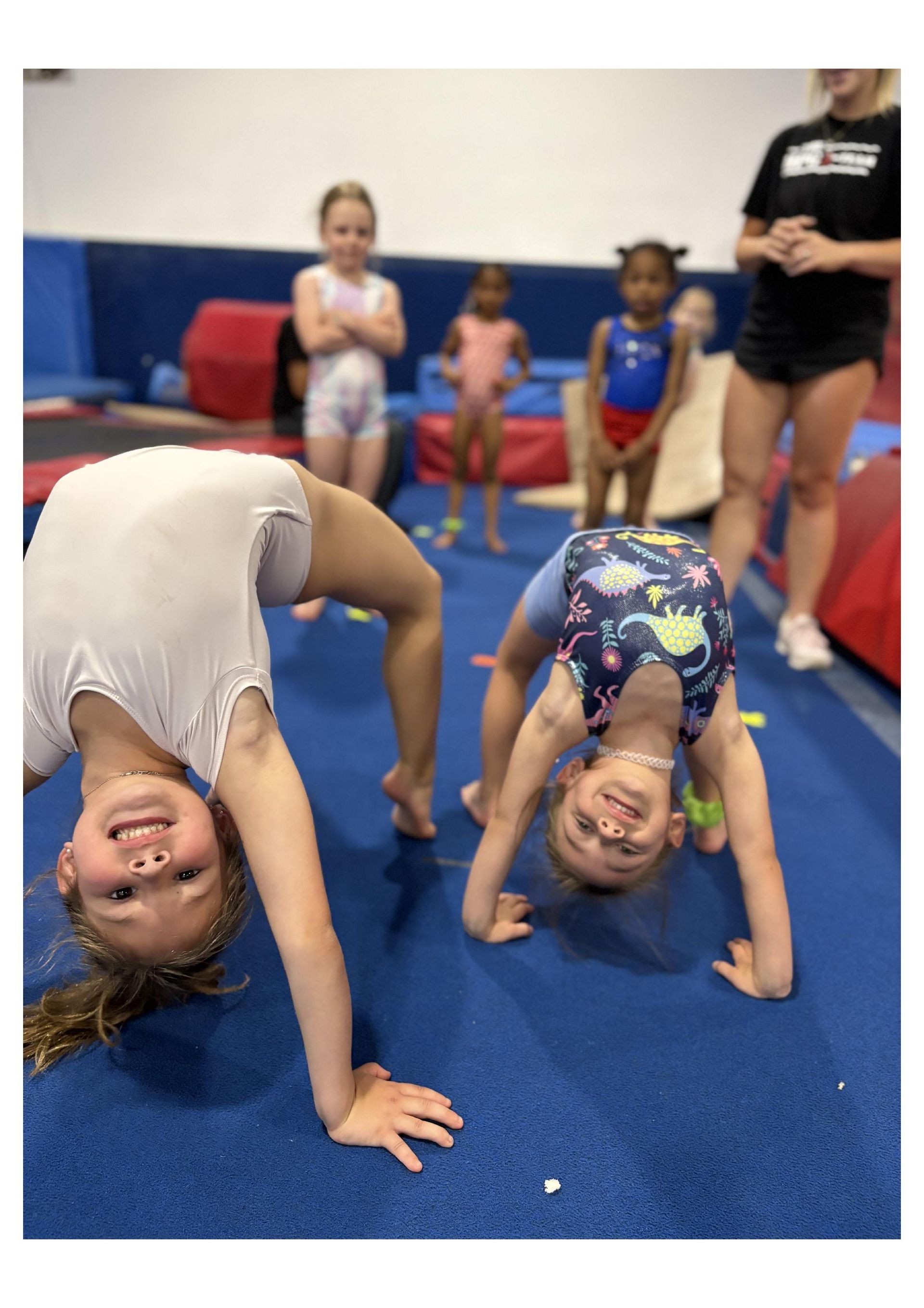 Two young girls are doing a handstand on a blue mat in a gym.
