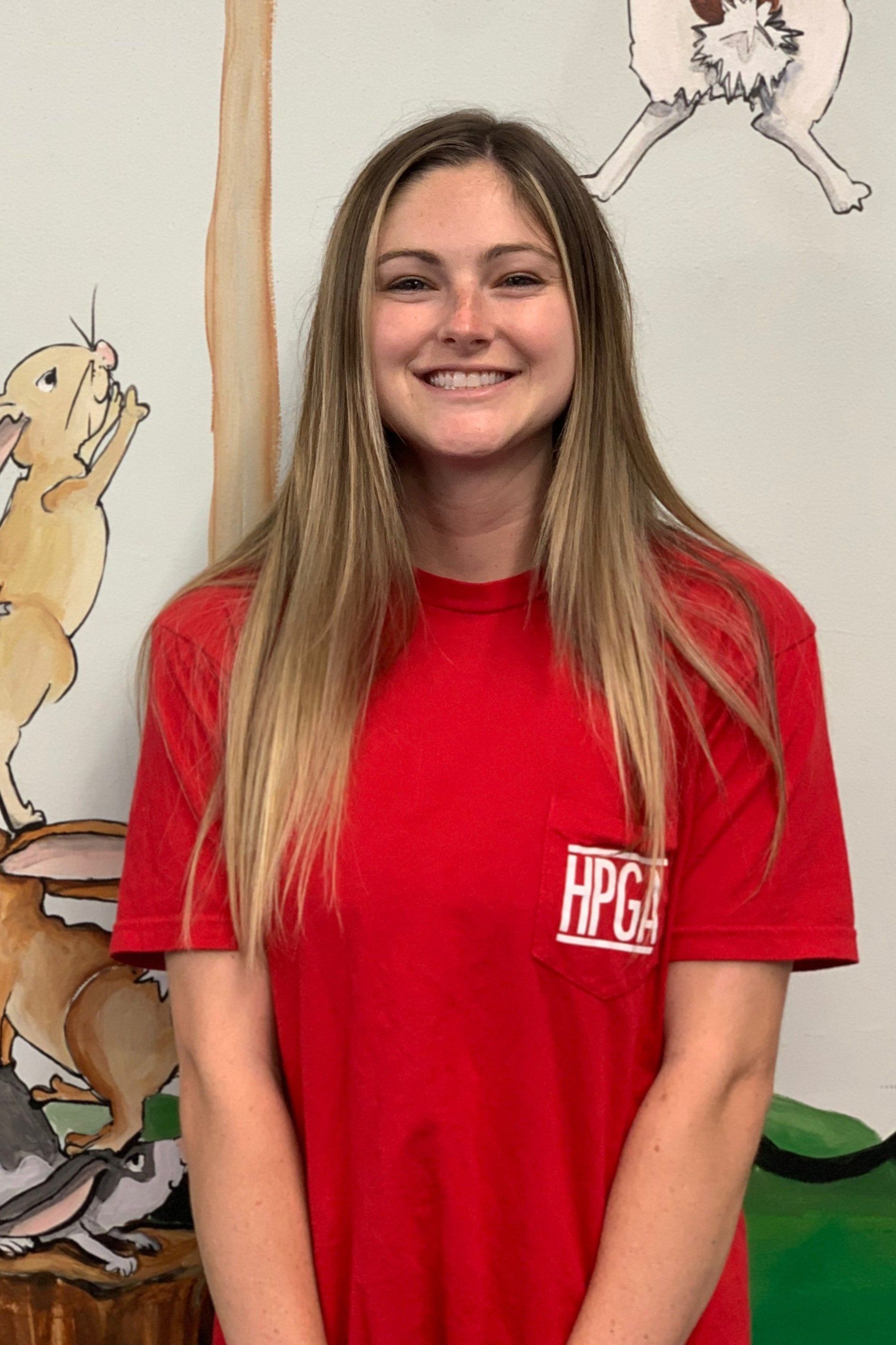 A woman in a red shirt is smiling in front of a painting of animals.