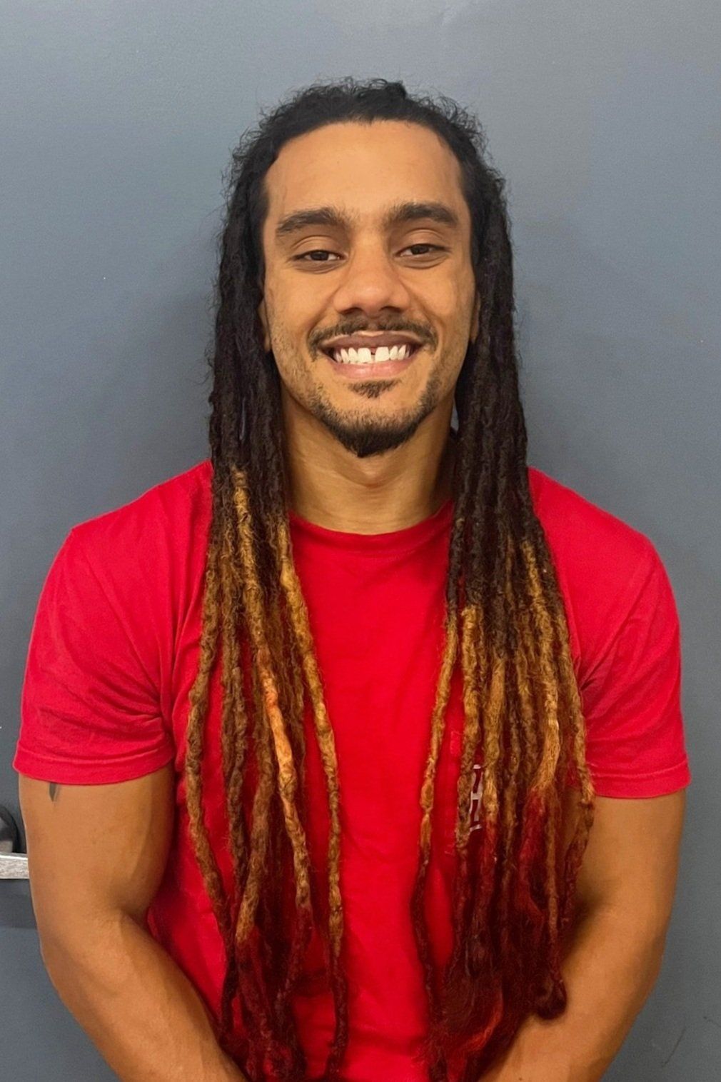 A man with dreadlocks is wearing a red shirt and smiling.
