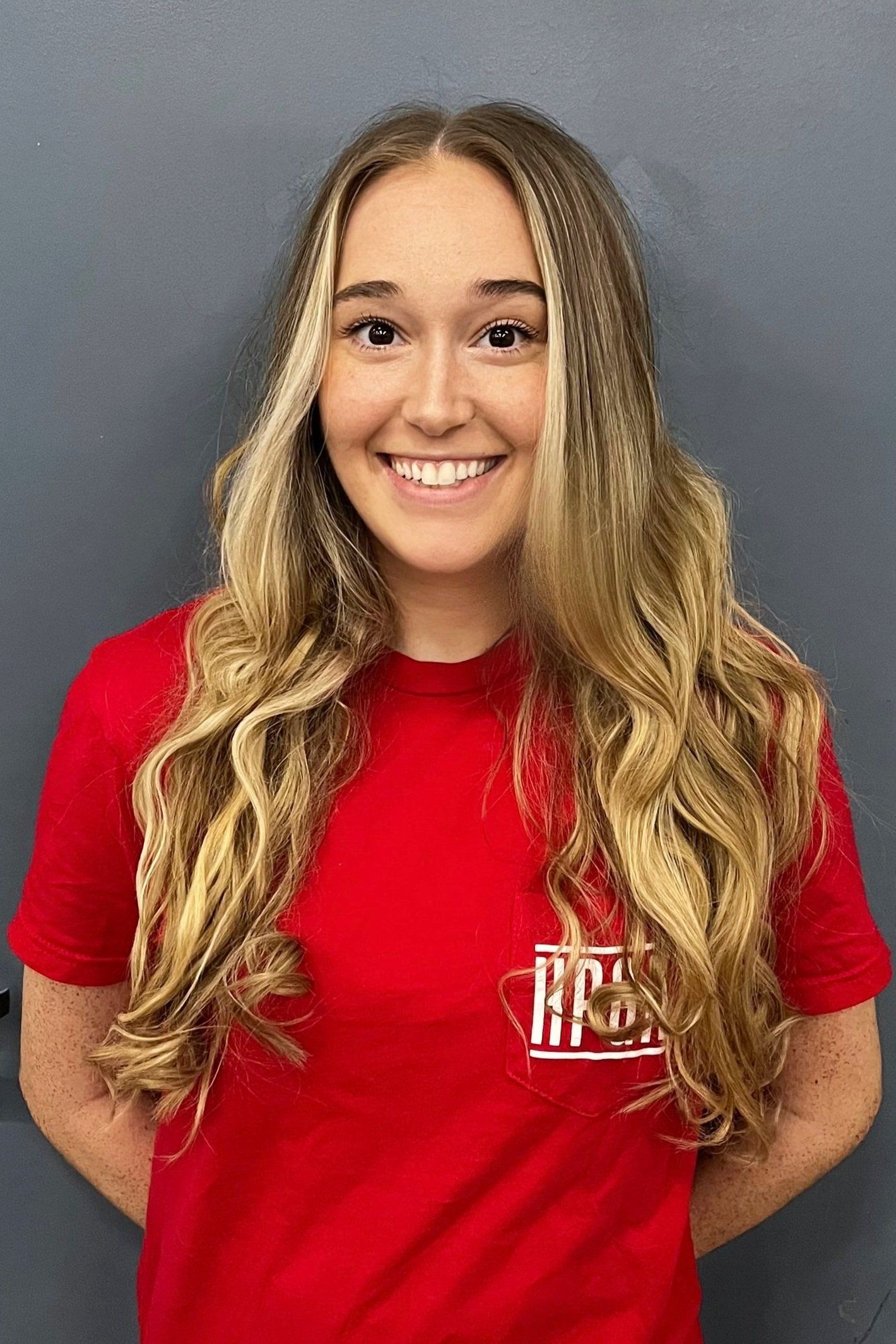 A woman with long blonde hair is wearing a red t-shirt and smiling.