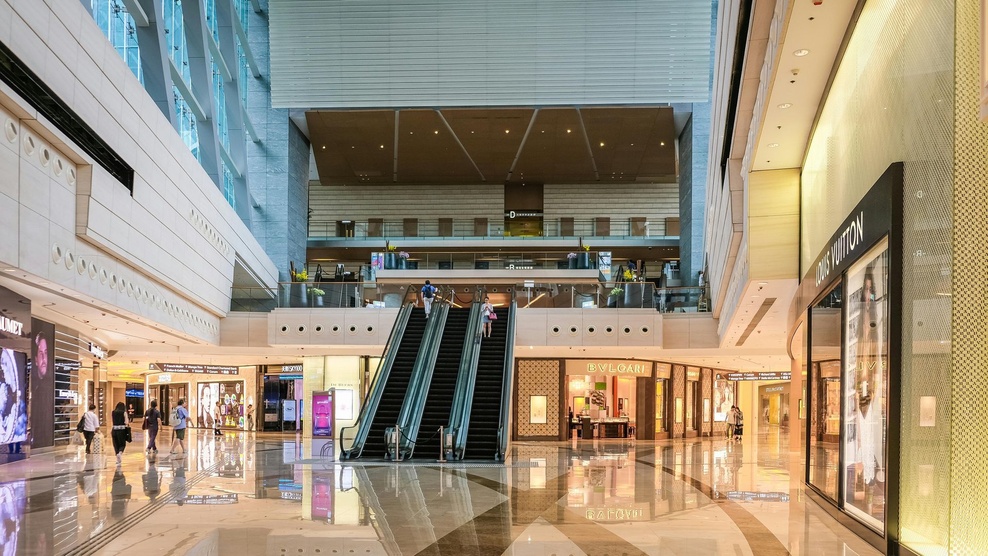 Interior of a modern shopping mall with escalators and multiple storefronts. People are seen walking and shopping.