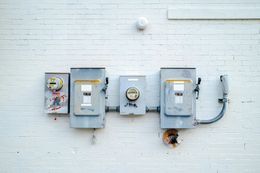 Electrical meters and boxes mounted on a white brick wall. They are weathered and gray, with some damage and wear.