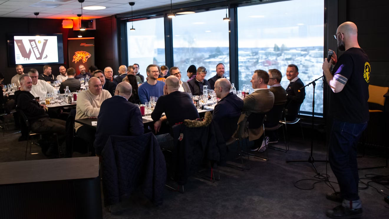 en mann står foran en mikrofon foran en gruppe mennesker som sitter ved bordene.