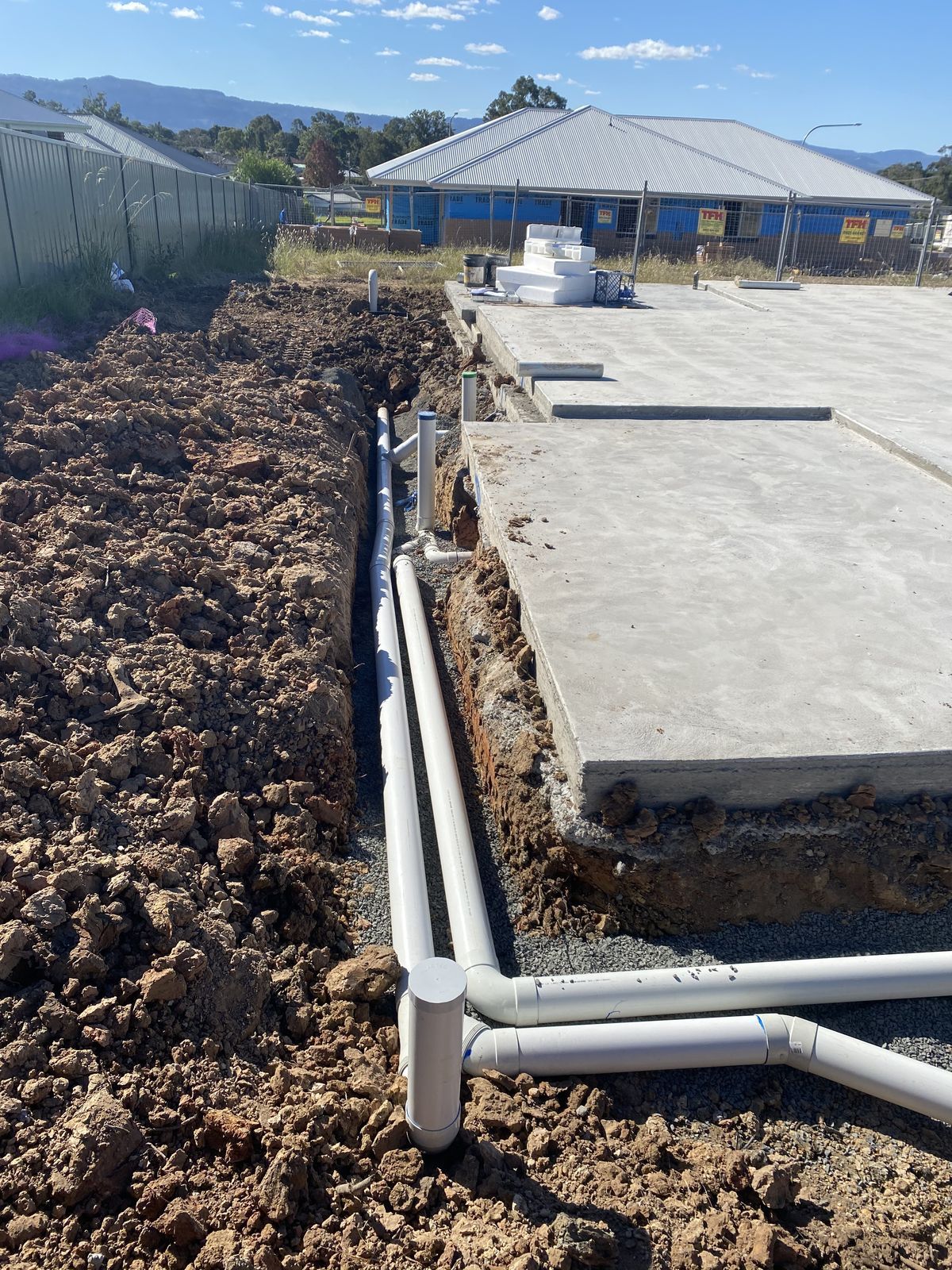 White plumbing pipes installed in a trench along the edge of a new concrete building foundation at a construction site.