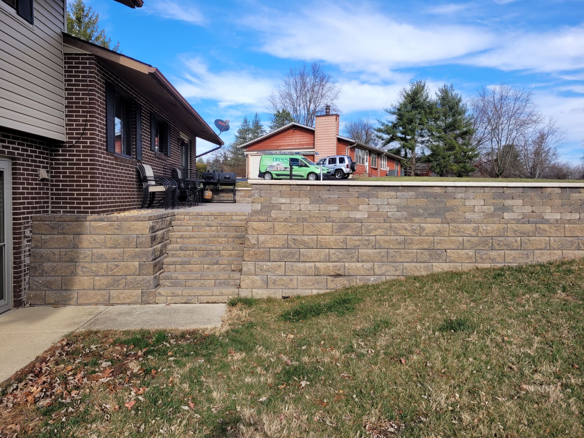 A brick house with a multi-level retaining wall and stone steps leading to a patio, set against a lawn and blue sky.