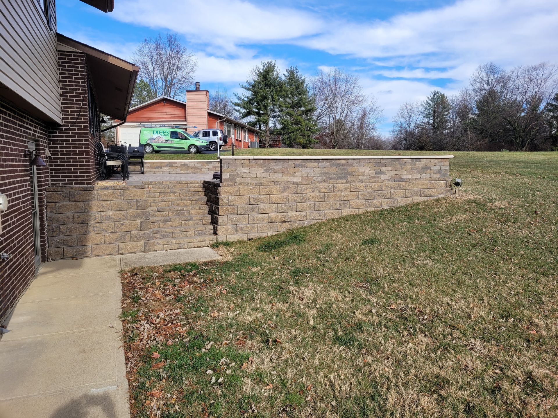 A tan, block-constructed retaining wall and patio area attached to the side of a brick house with a grassy lawn.