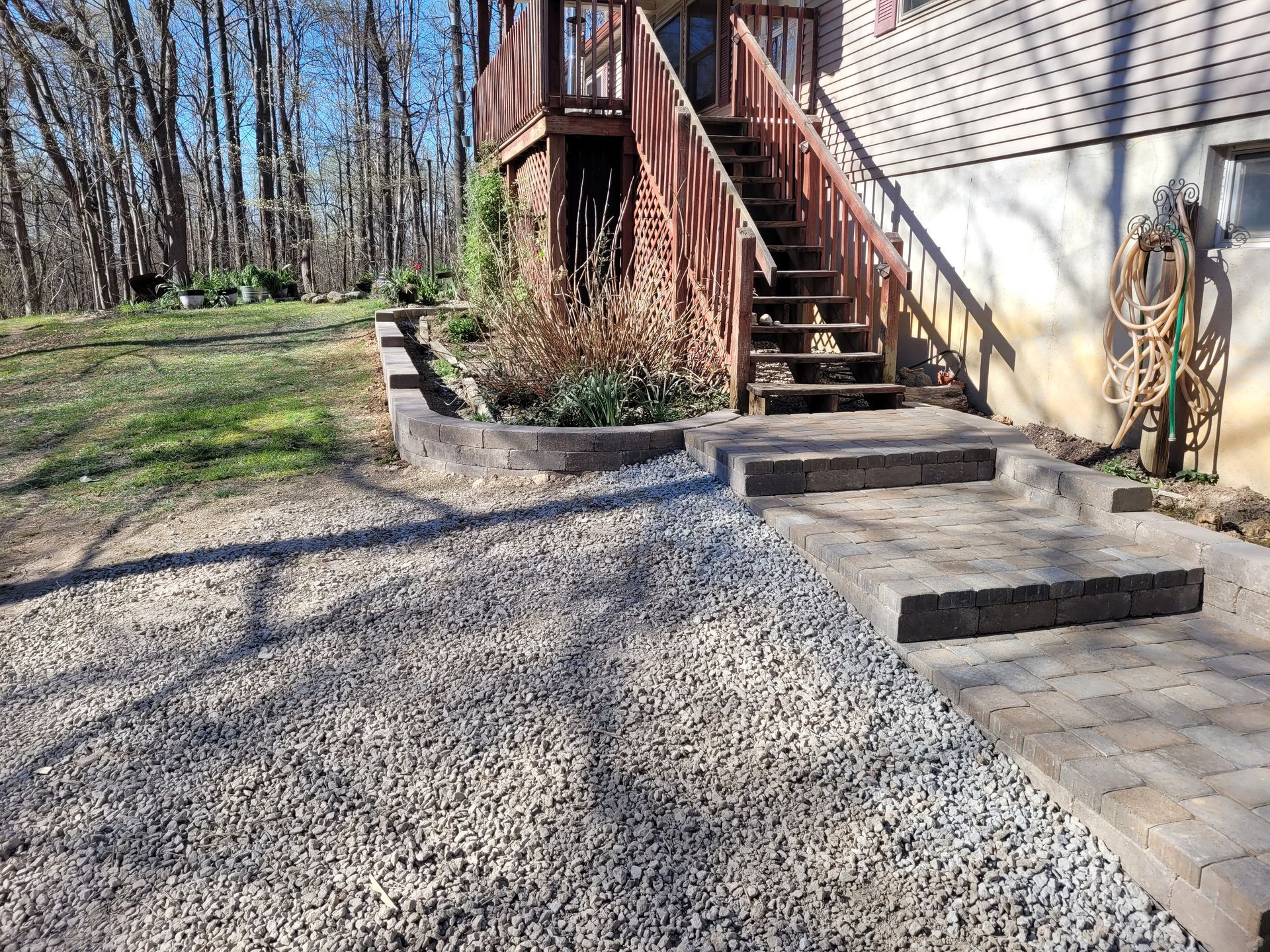 A gravel path leading to stone steps and a wooden deck attached to the side of a house, set against a background of trees.