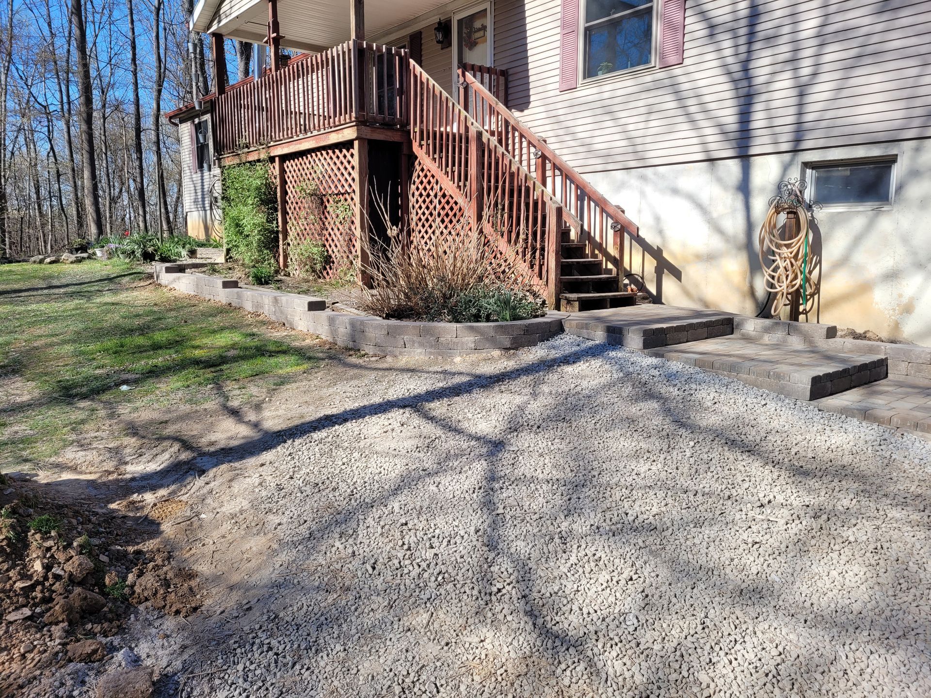 A pile of fresh gravel sits in front of a house with wooden stairs and a retaining wall, showing a landscaping project.