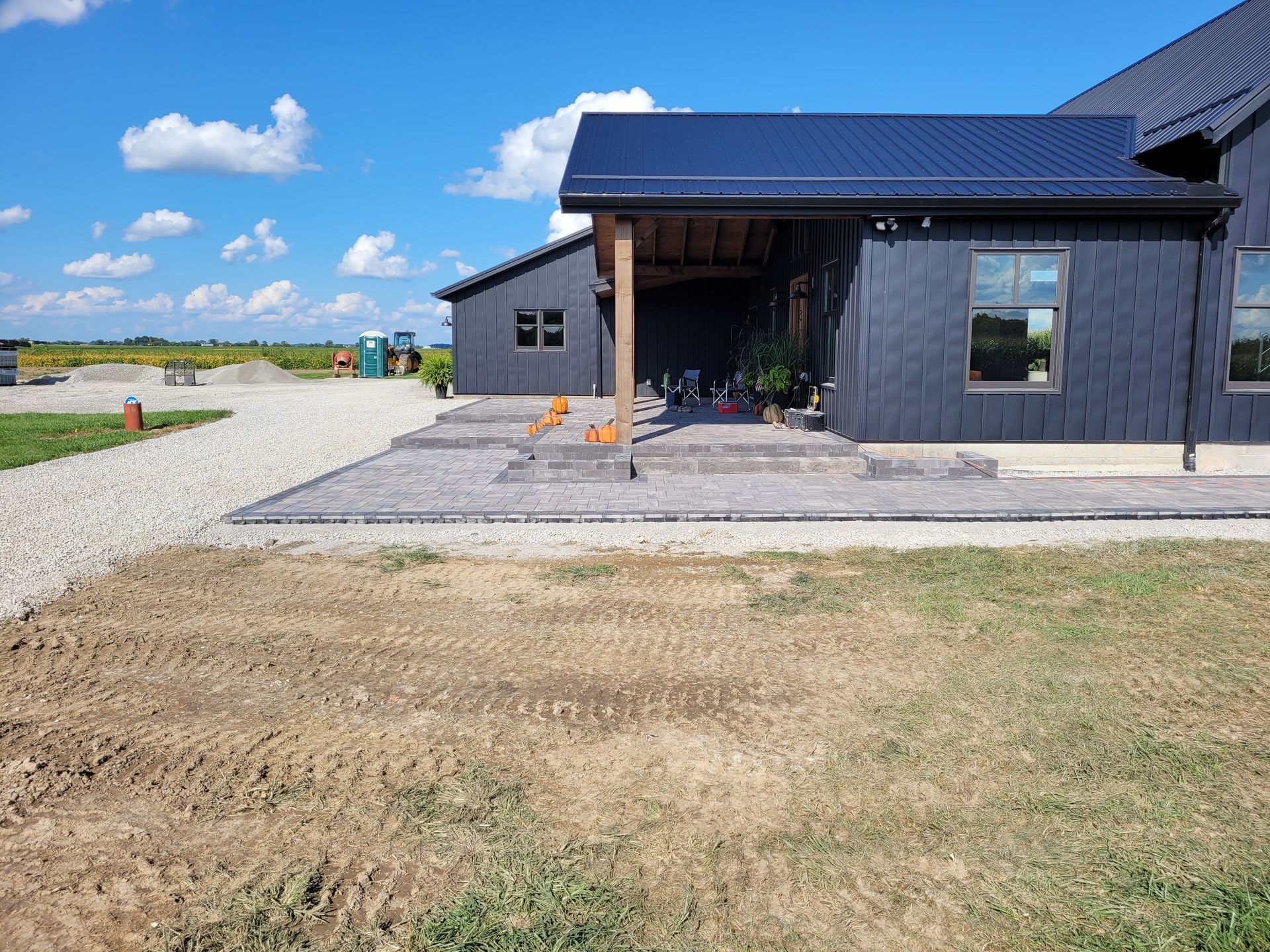 A modern dark-sided house with a paved patio and covered entryway under a clear blue sky, next to a gravel driveway.