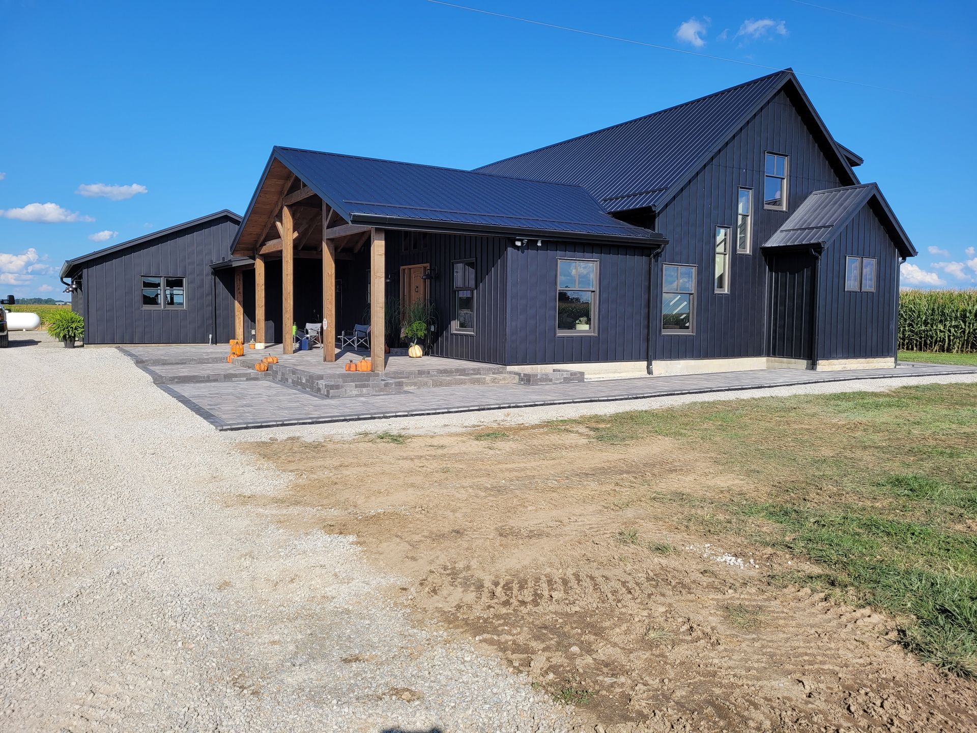 A black, modern farmhouse with a wooden porch and metal roof, situated on a gravel driveway under a bright blue sky.