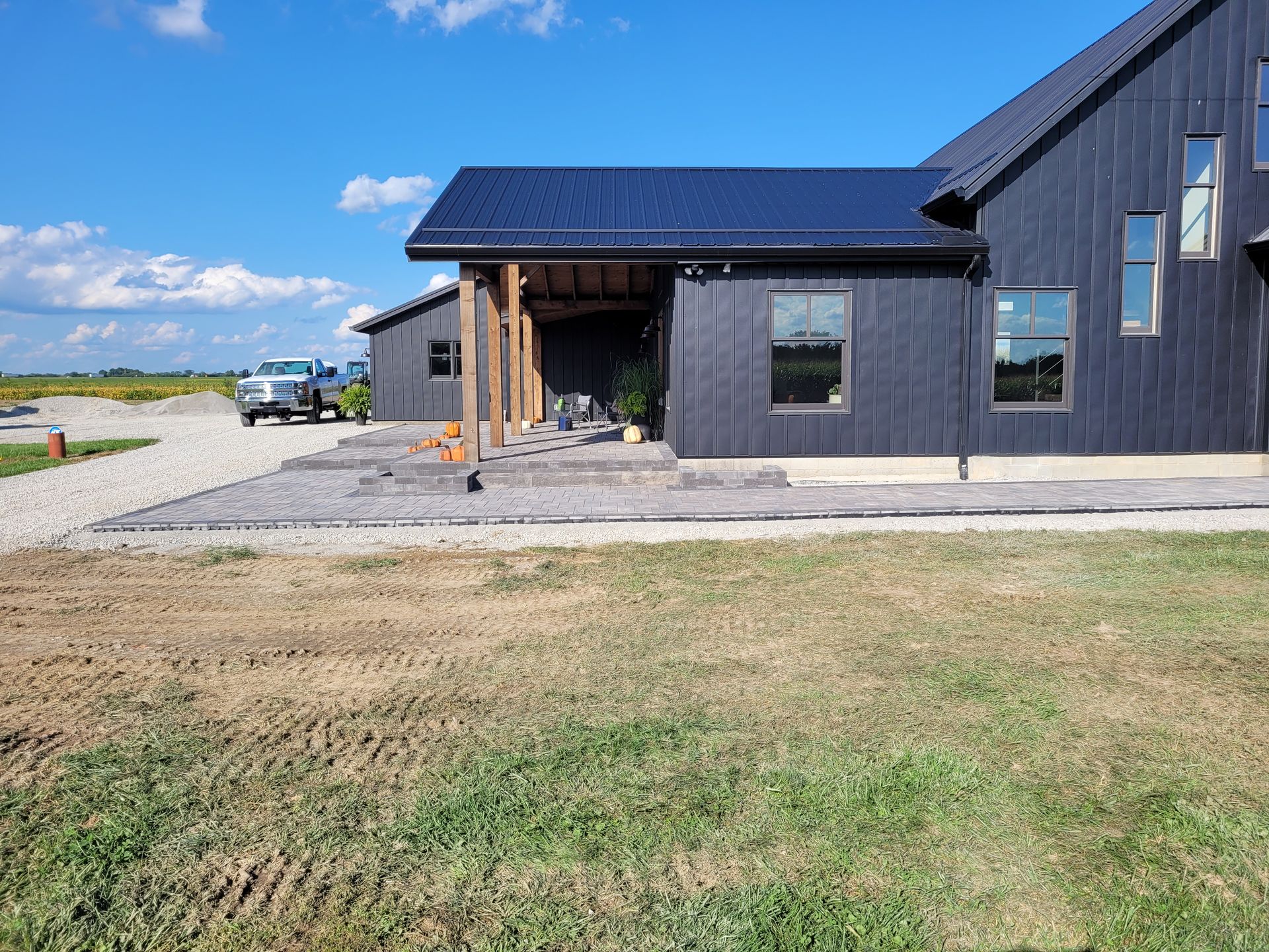 A black, modern farmhouse with a covered patio, gravel driveway, and unfinished lawn under a blue sky.