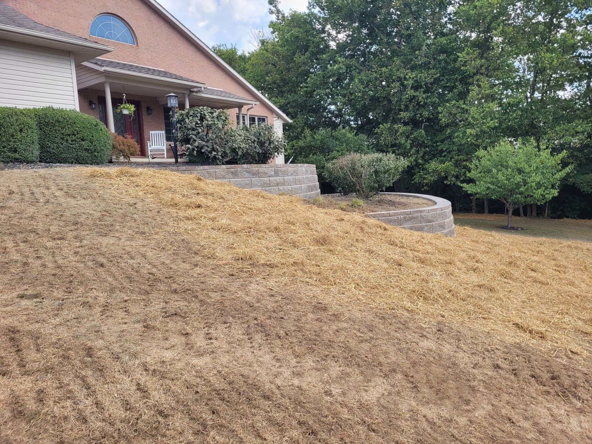 A brick house sits atop a tiered landscape with a freshly seeded, straw-covered slope in the foreground.