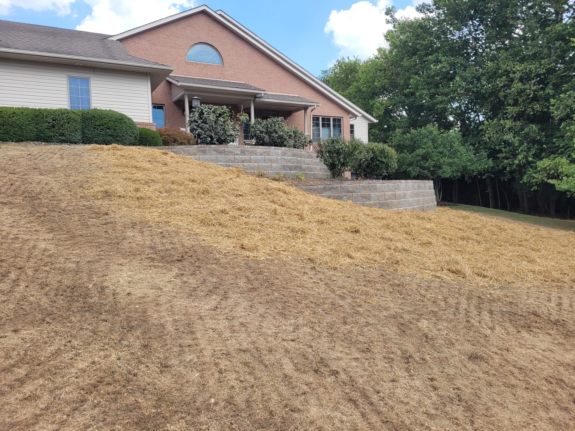 A brick and siding house with a tiered stone retaining wall overlooking a large, straw-covered sloped lawn.