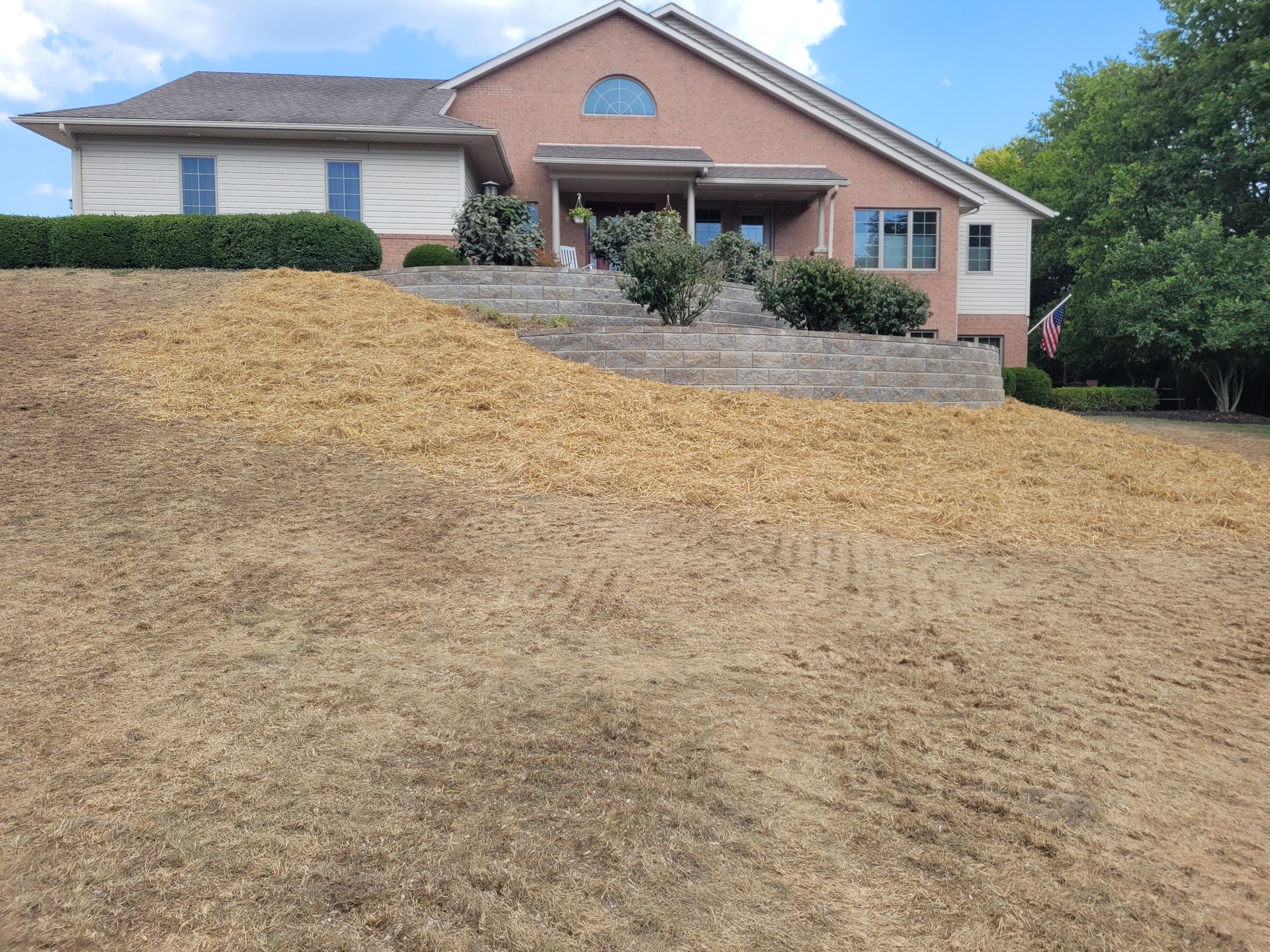 A tan-colored house with a stone retaining wall and a sloped front yard recently covered with straw for grass seeding.