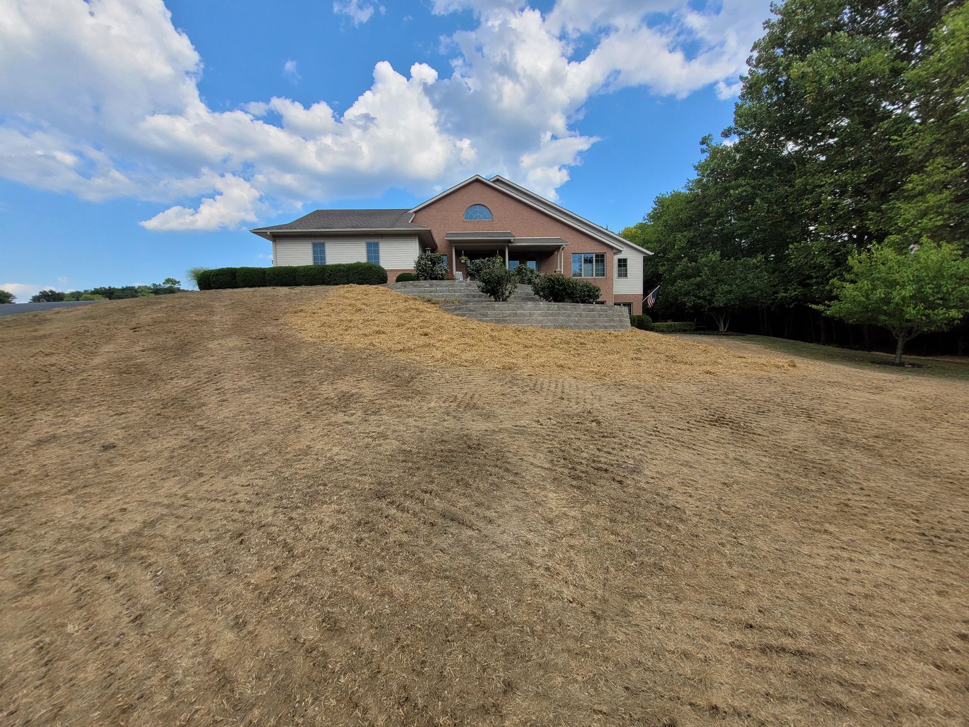 A house with beige and brick siding sits atop a large, sloped hill of dry, brown grass under a bright, partly cloudy sky.