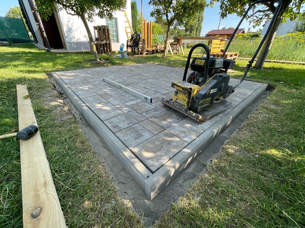 A vibrating plate compactor sits on a newly laid patio of grey stone pavers in a grassy yard.