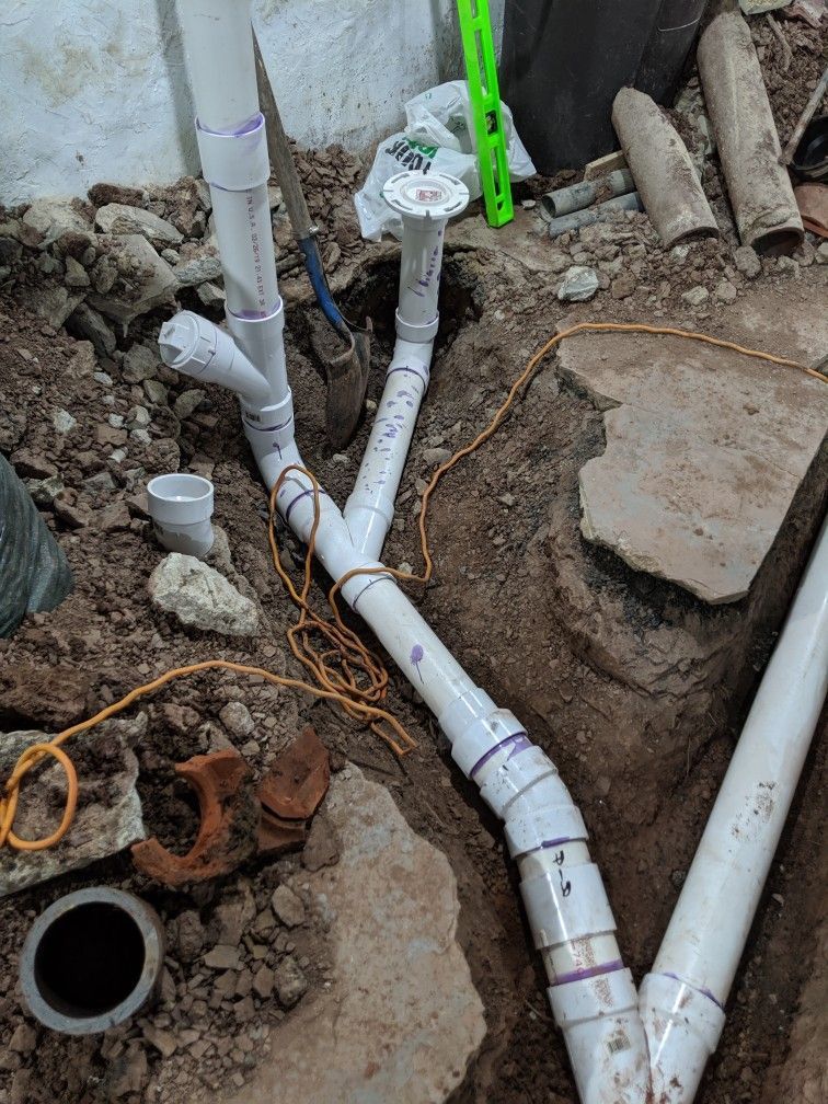 A trench filled with dirt reveals newly installed white PVC drainage pipes connected with various fittings and a cleanout.