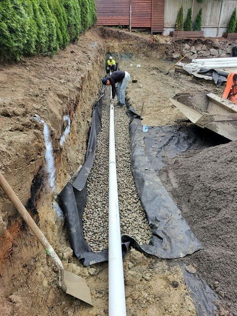 A worker installs a white perforated drain pipe in a gravel-filled trench lined with black landscape fabric.