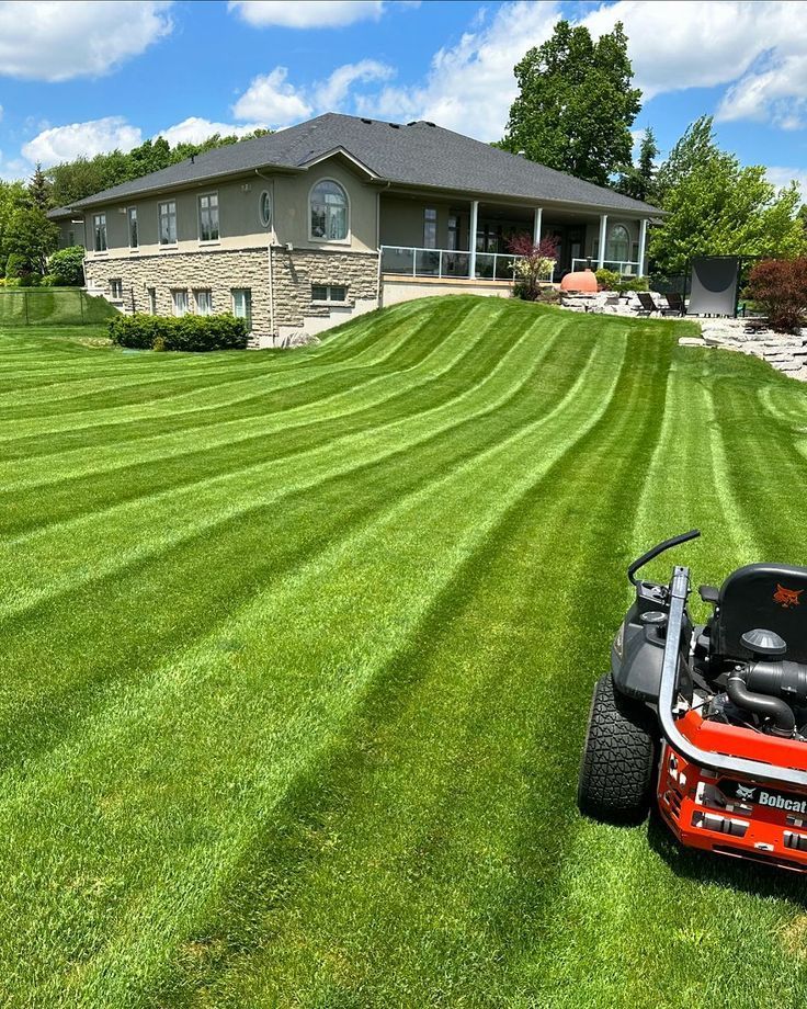A bright red zero-turn mower sits on a lush green lawn with crisp, professionally mowed stripes, leading to a large house.