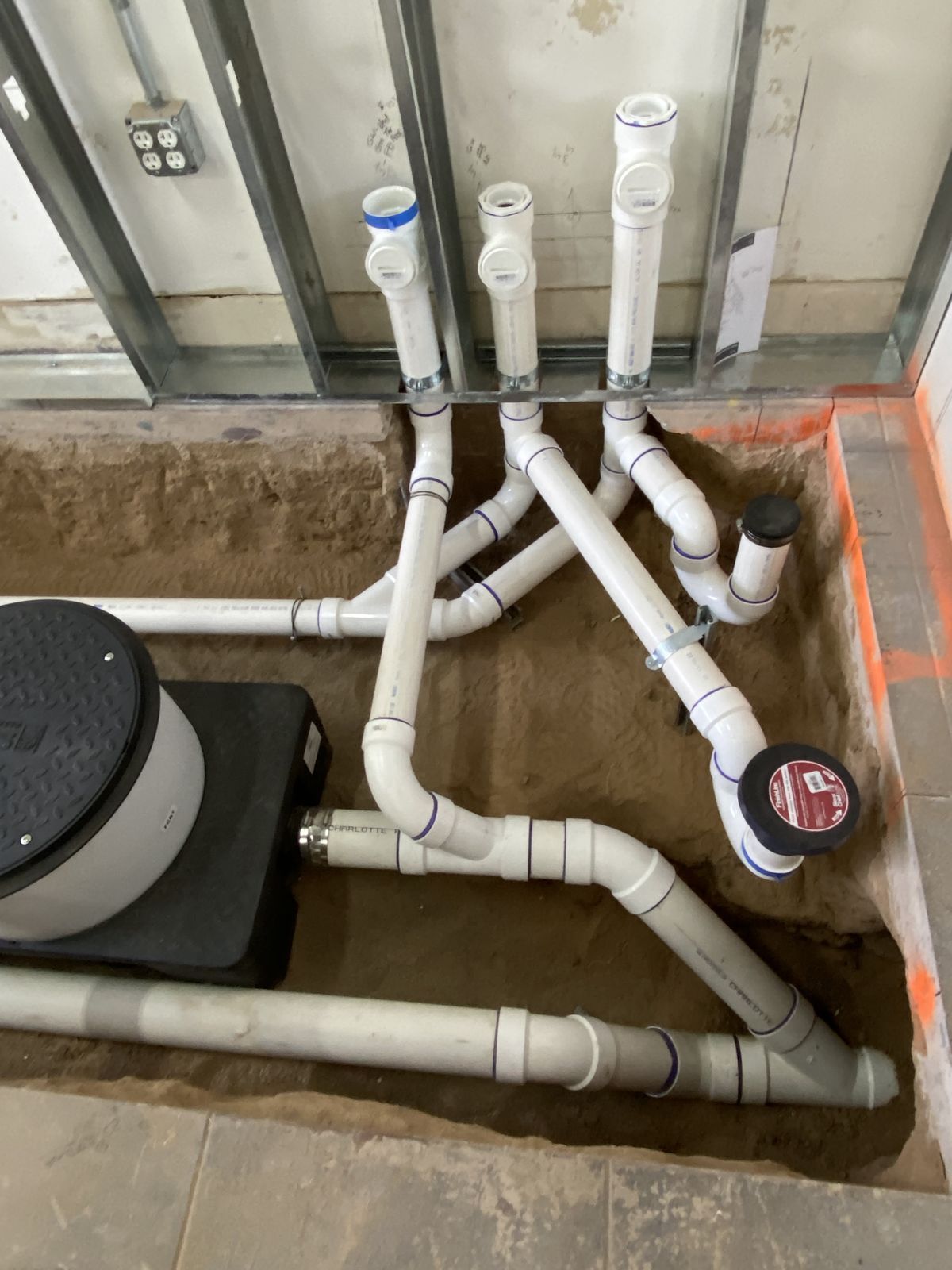 PVC plumbing pipes installed in a trench near a black sump pump pit within a stud-framed wall construction site.