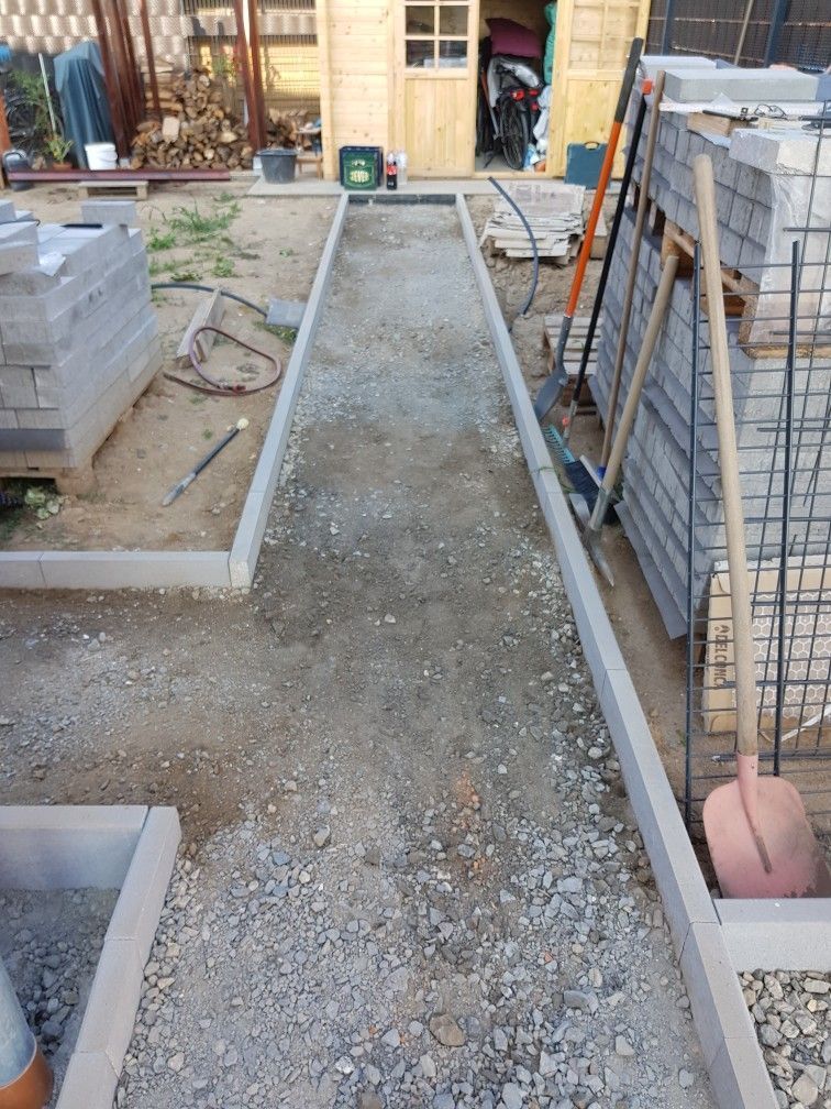 A narrow gravel walkway framed by concrete edgings in an outdoor construction site, with stacked paving materials nearby.