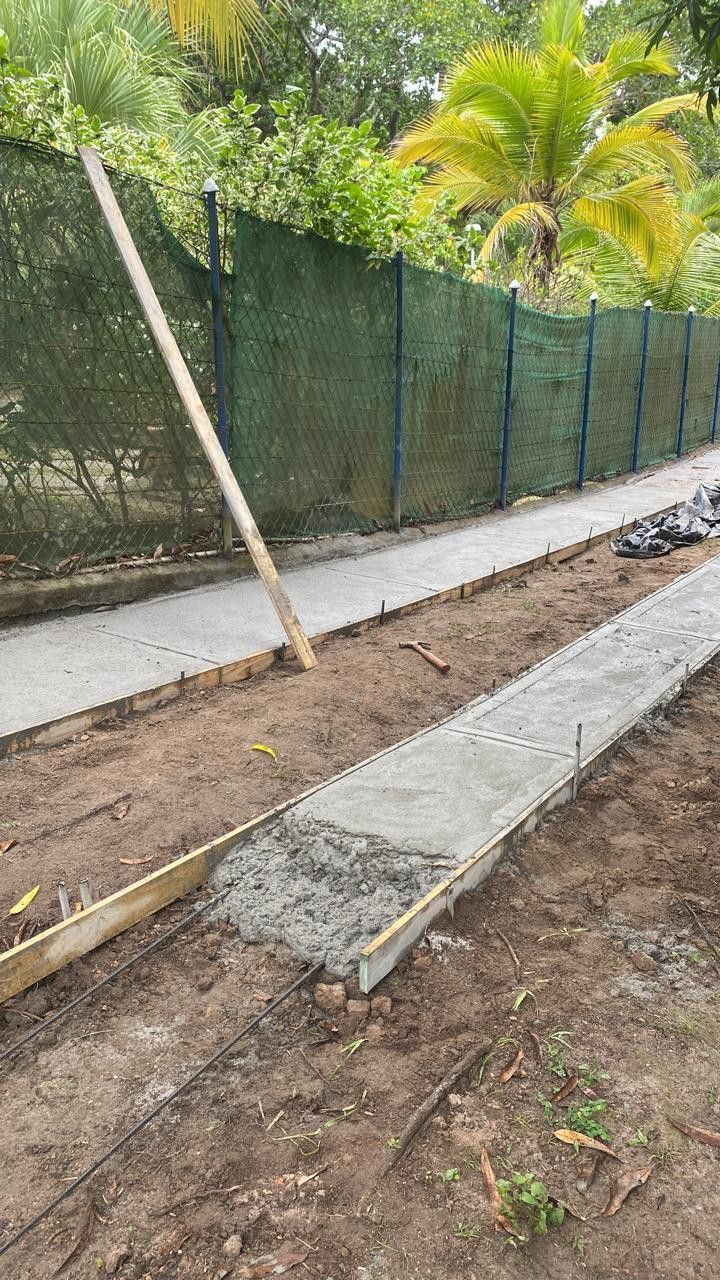 A backyard construction scene with a finished concrete sidewalk and a new narrow concrete form being poured near a fence.