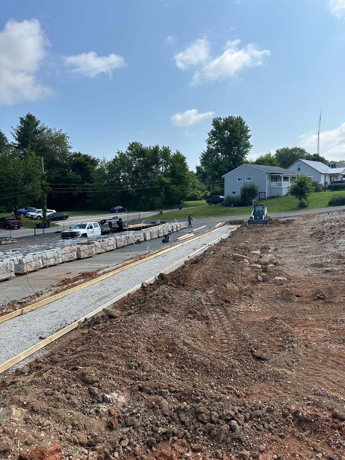 A construction site showing a gravel path, a row of concrete blocks, a white truck, and nearby houses under a blue sky.