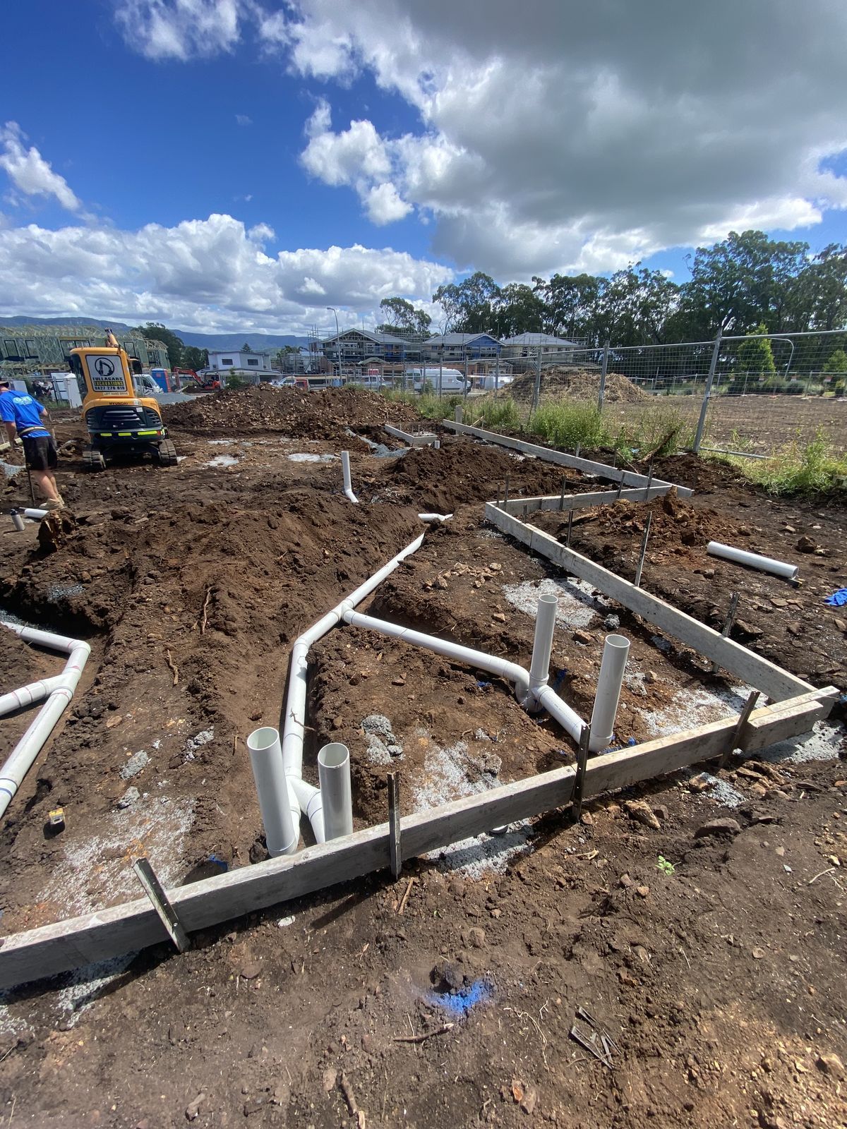 Construction site with trenches, concrete formwork, and white plumbing pipes installed in soil under a cloudy sky.