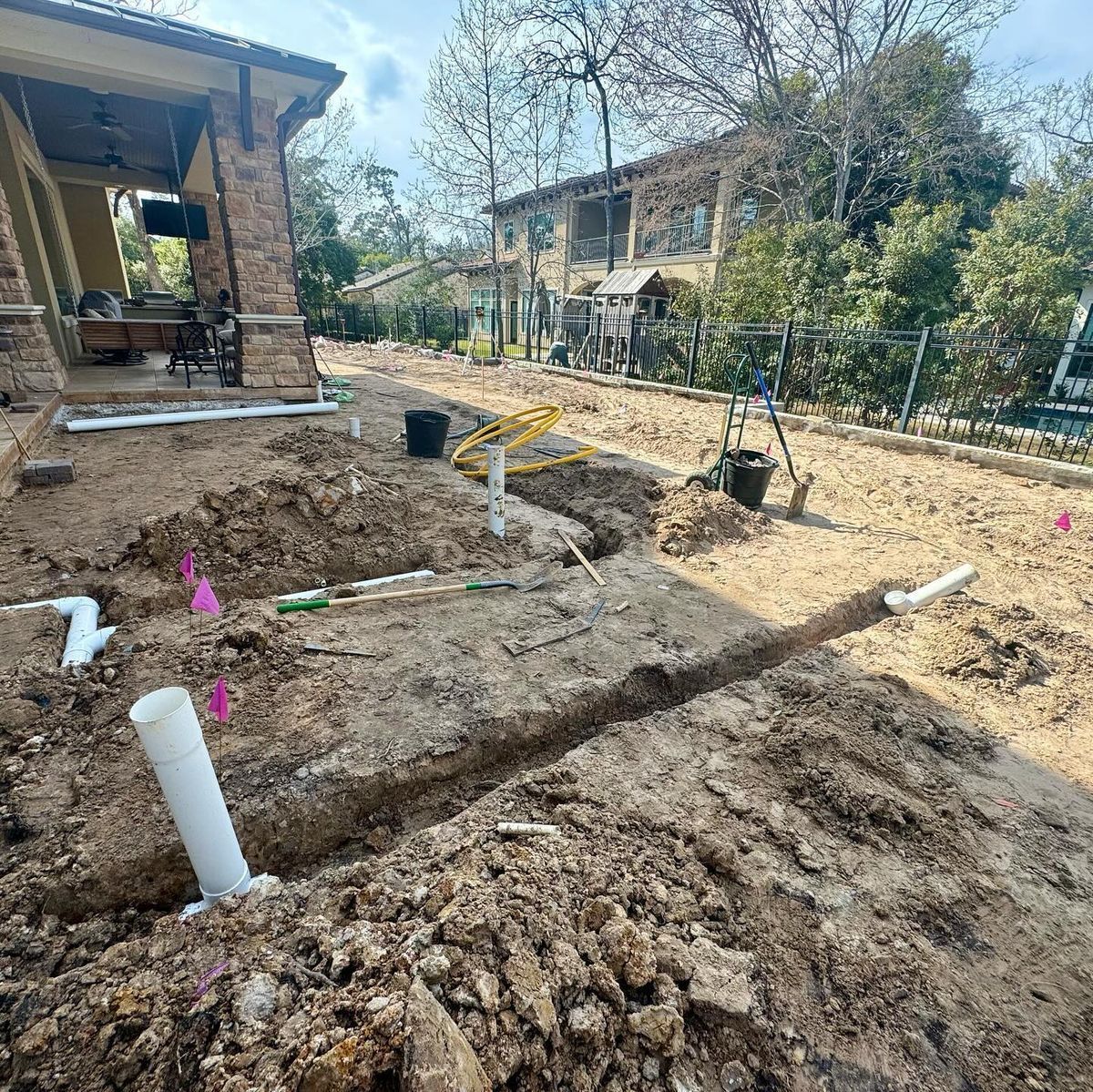 A backyard construction site shows trenches and white PVC pipes laid in the dirt near a residential patio.