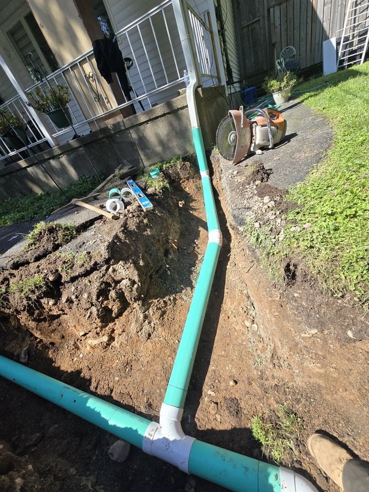 A green drainpipe assembly installed in a trench next to a residential foundation and lawn.