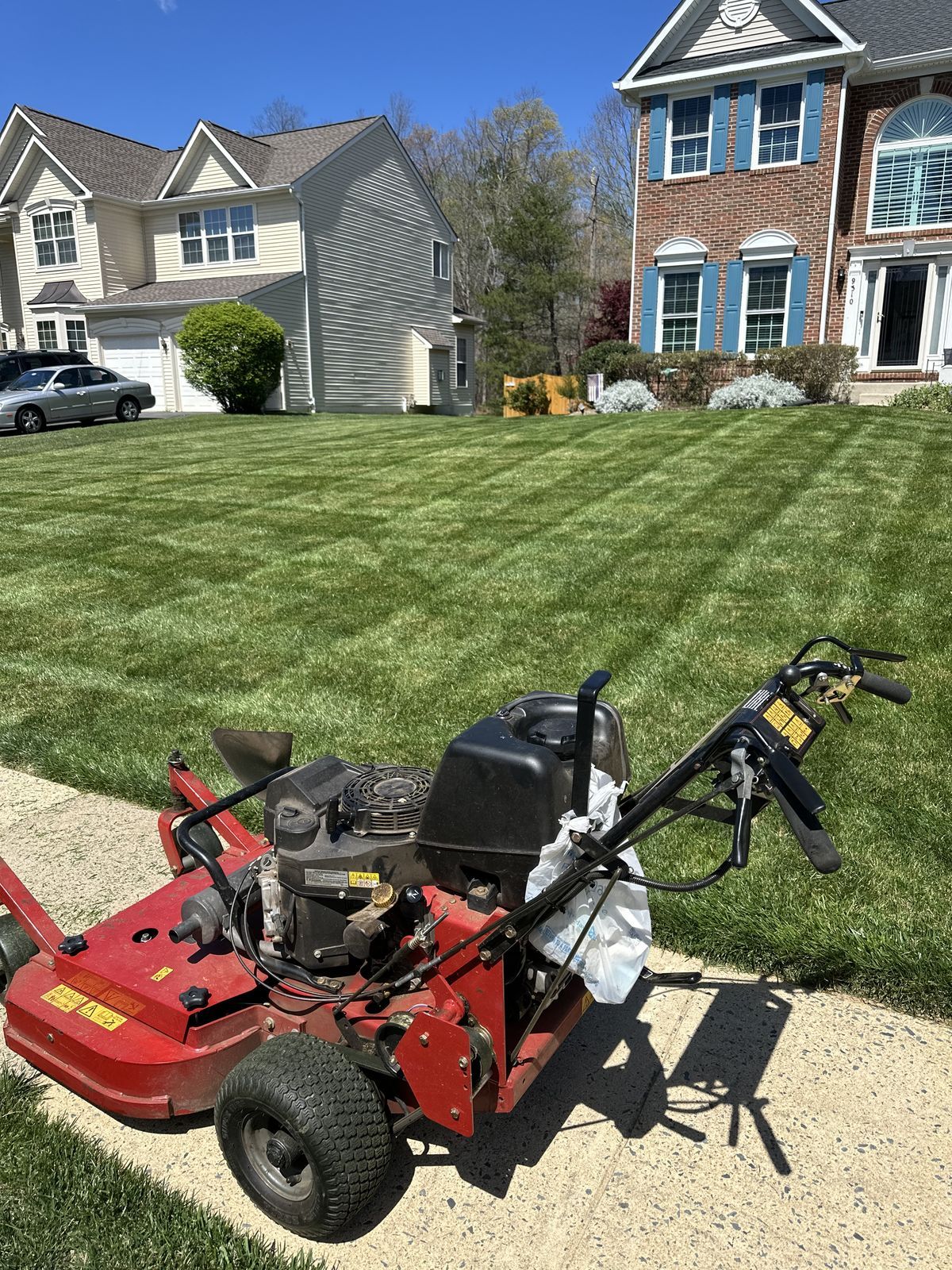 A red commercial walk-behind lawn mower sits on a sidewalk next to a freshly mowed lawn with prominent striping patterns.