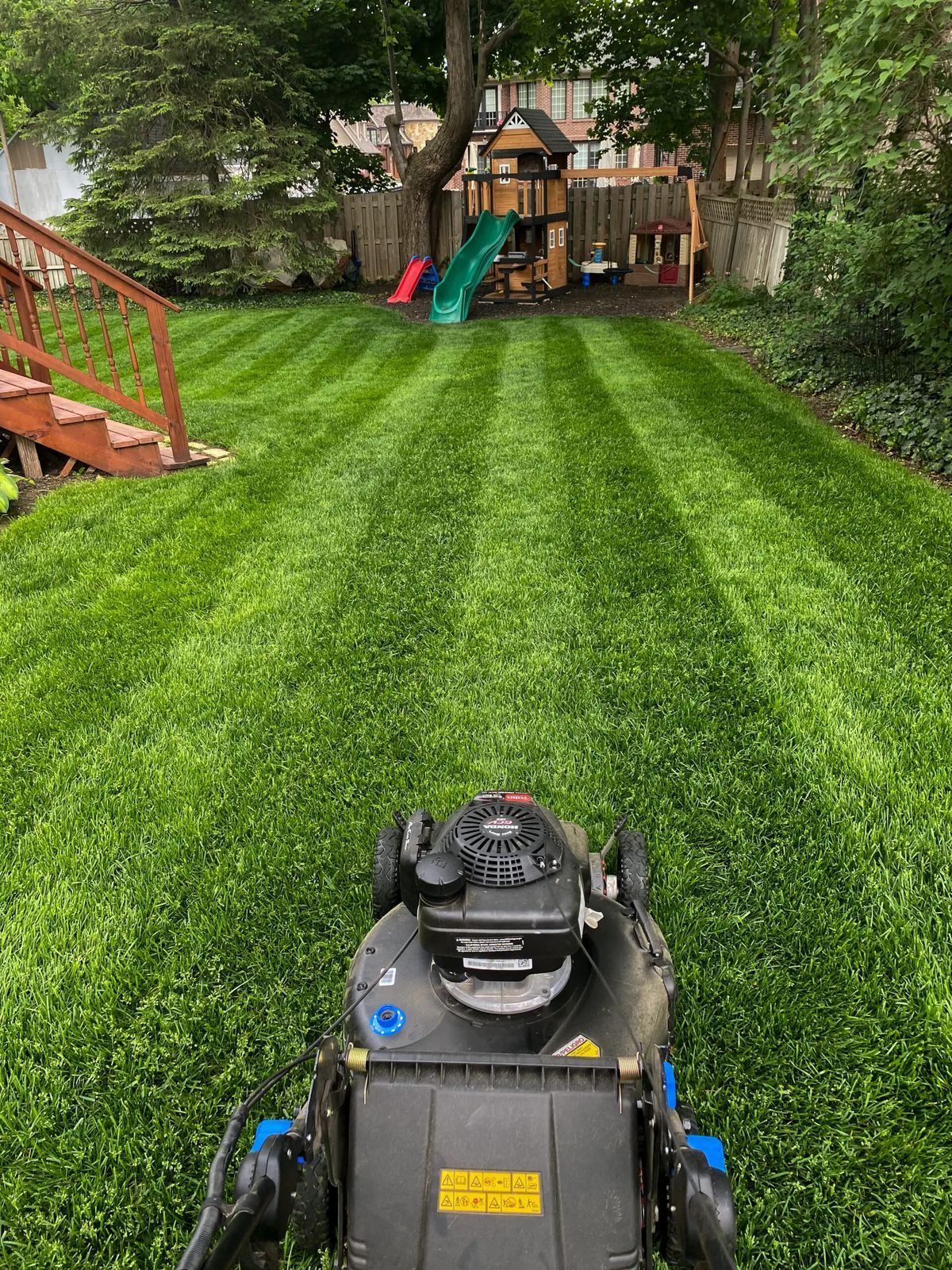 A first-person view from a lawnmower pushing through a lush green backyard with neatly striped grass and a playground.