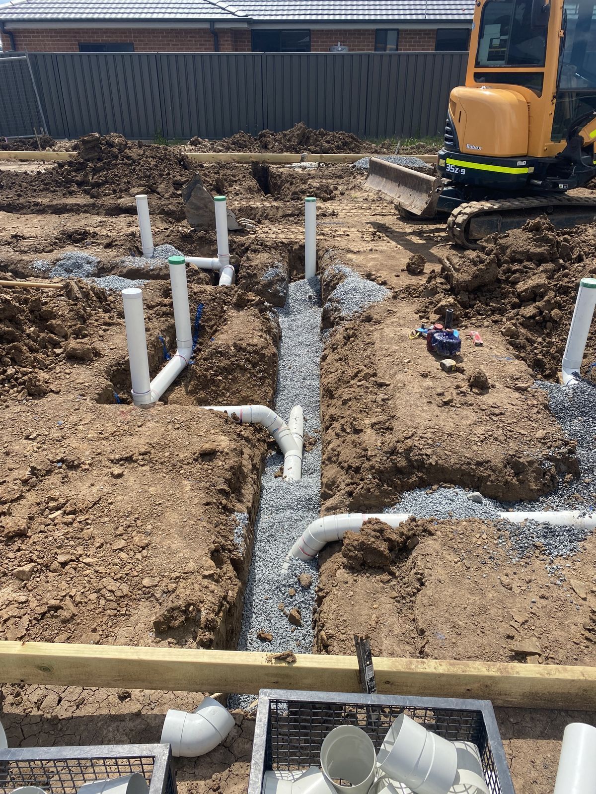 Construction site showing white PVC plumbing pipes installed in gravel-filled trenches with an excavator in the background.