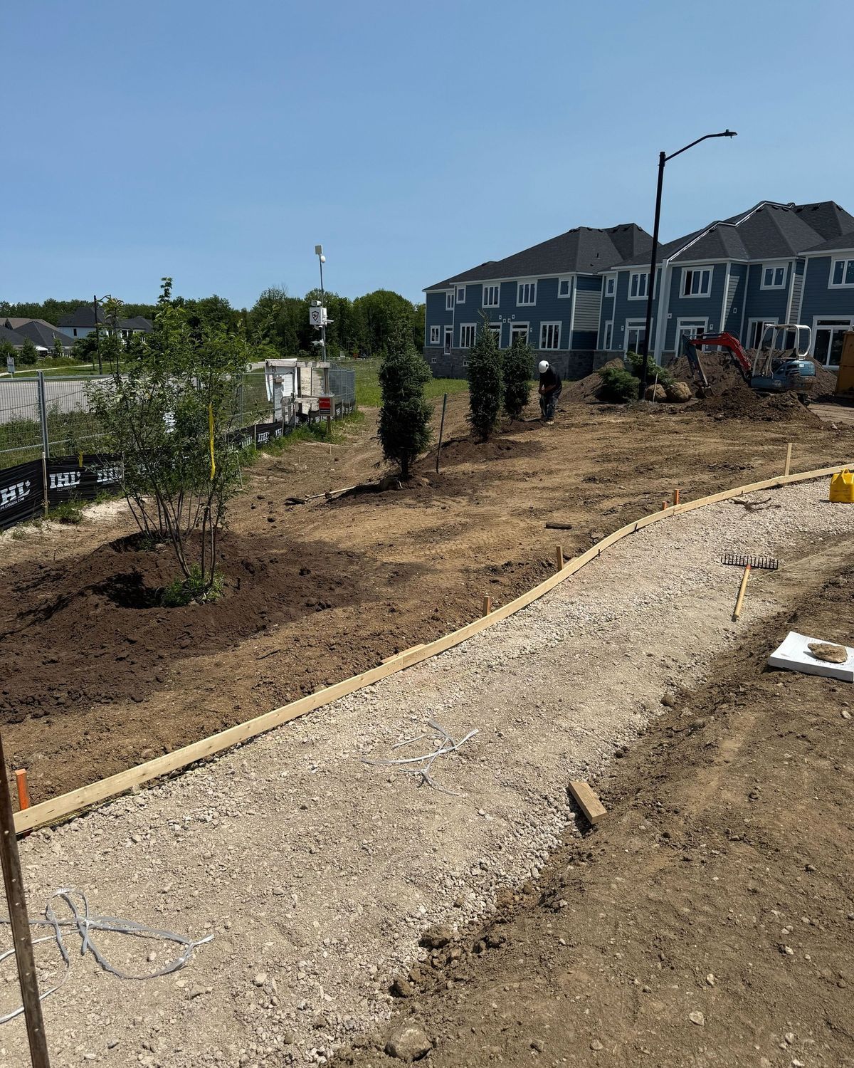 A gravel path under construction with planted trees and shrubs in front of blue suburban townhouses on a sunny day.