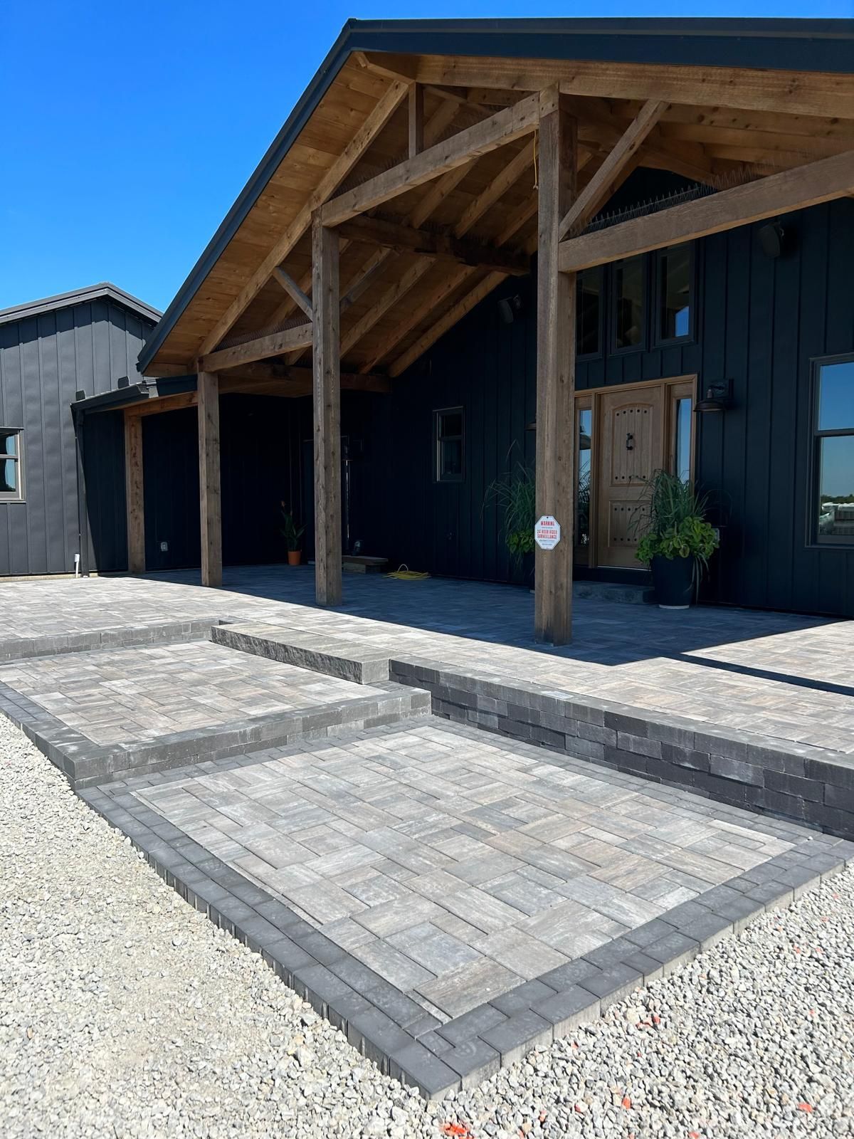 A dark, modern house with wooden timber framing over a patio entrance, featuring stone paver steps and a gravel foreground.