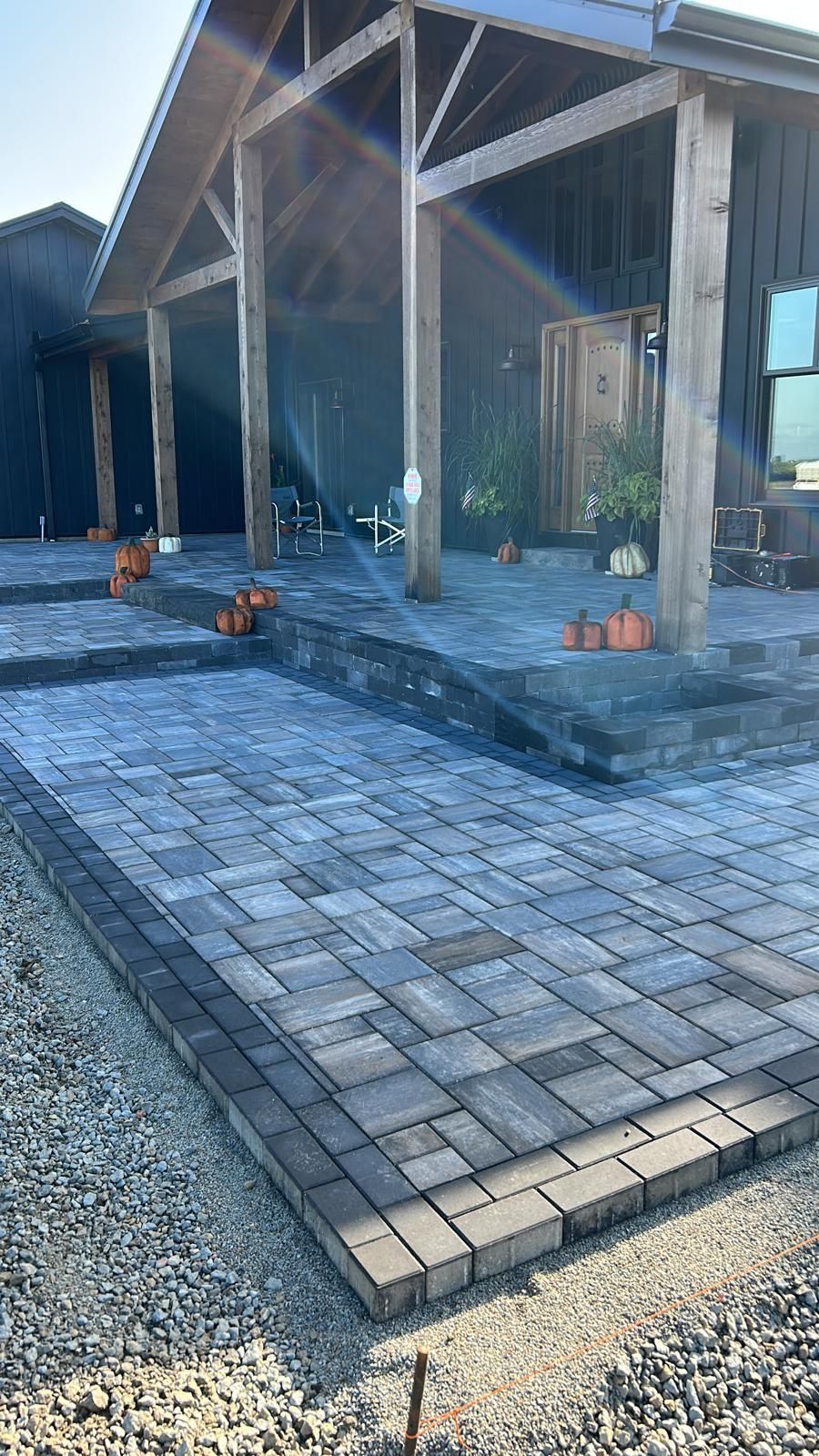 Modern dark-sided house with a large stone paver patio, wooden porch pillars, and decorative pumpkins on the steps.