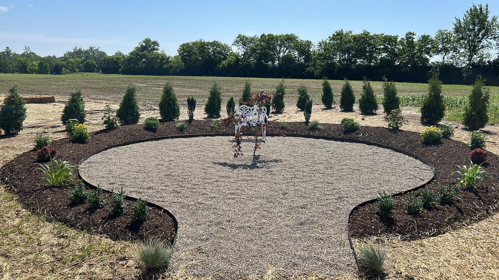 A circular gravel garden bed surrounded by dark mulch, small green shrubs, and a row of evergreen trees in a field.