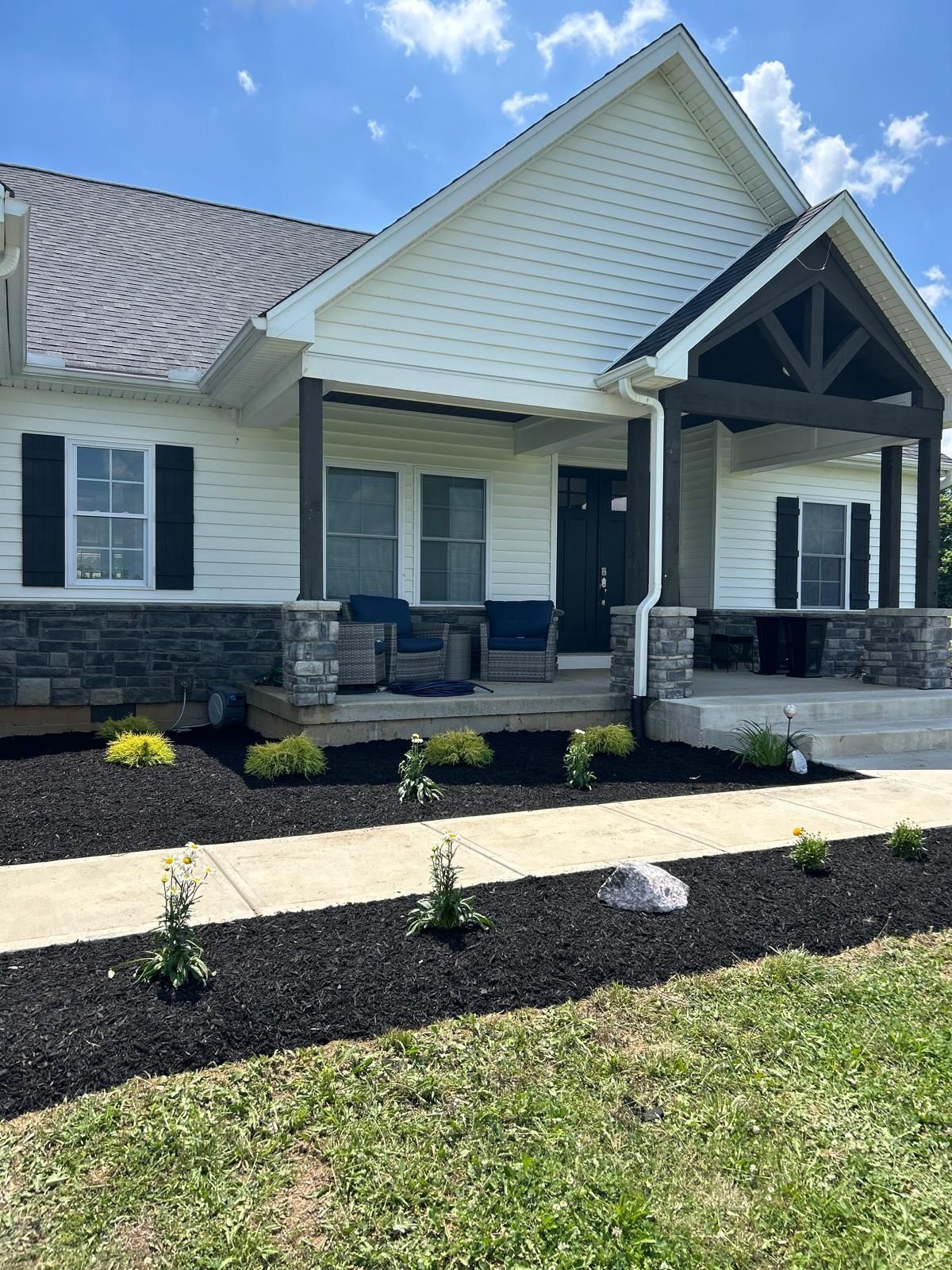 A cream-colored house with stone siding, a dark porch, a concrete walkway, and landscaped garden beds with black mulch.