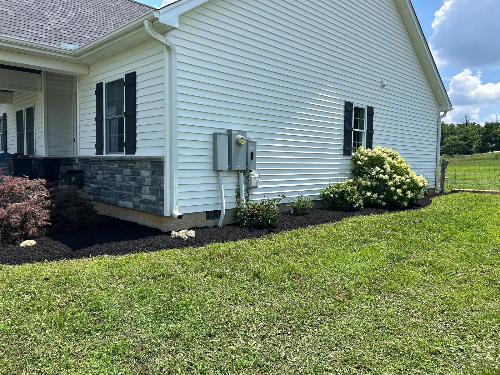 A side view of a white vinyl-sided house with a stone foundation, electrical boxes, and landscaping in a grassy yard.