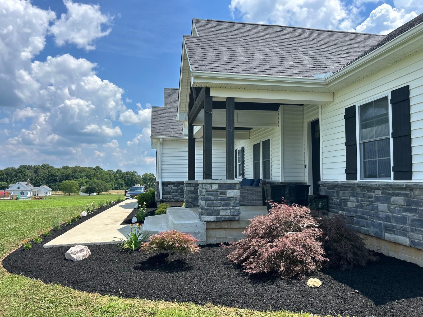 A cream-colored house with black shutters and a stone-based front porch under a bright blue sky with fluffy white clouds.
