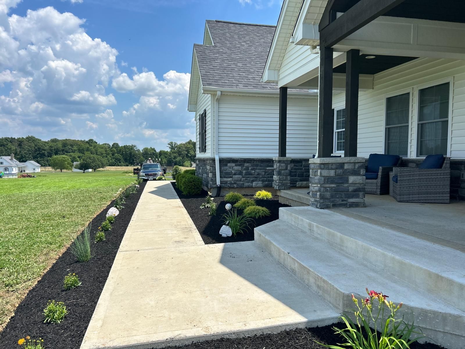 A concrete walkway leads to a white house porch with stone pillars, dark furniture, and mulch beds with small shrubs.